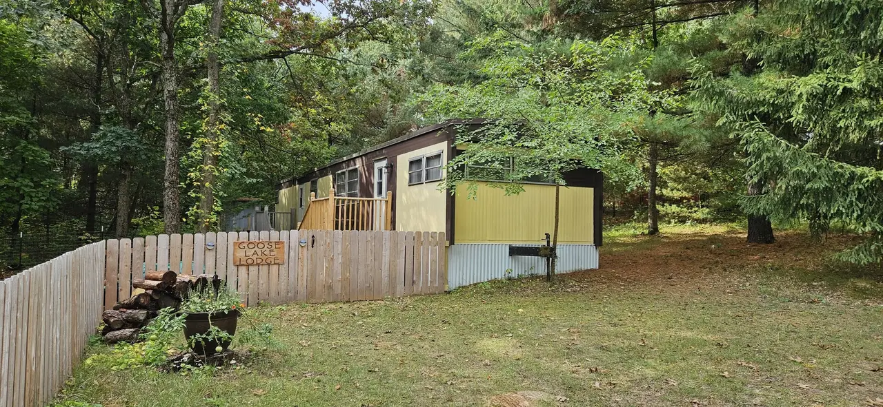 A mobile home with yellow and brown siding situated in a wooded area surrounded by green trees and a grassy yard, enclosed by a wooden fence with a sign that reads 'Goose Lake Lodge.' There is a woodpile in a planter and a grassy area in front of the home.
