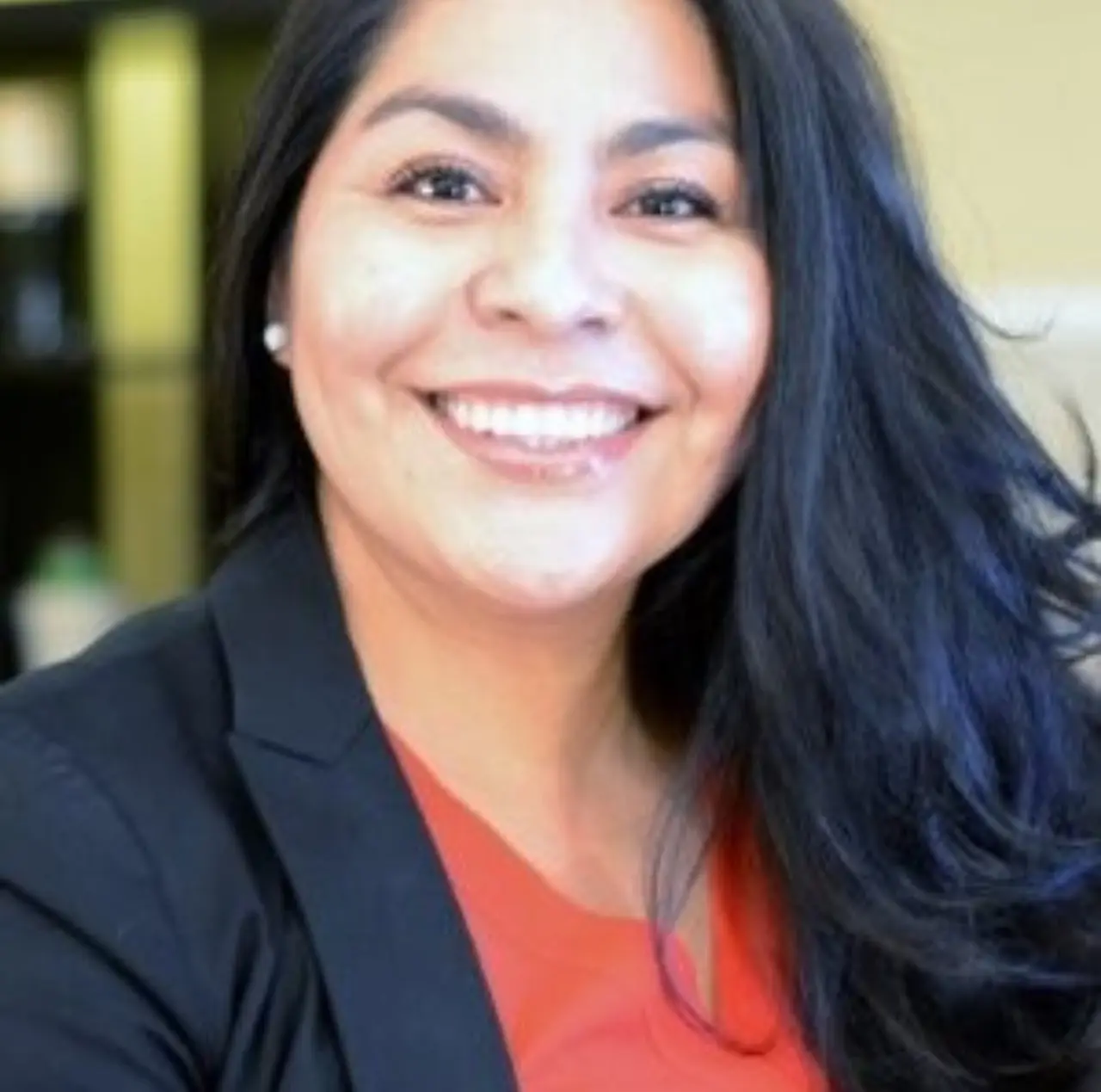 A smiling woman with long dark hair in a black blazer over an orange top, photographed indoors.