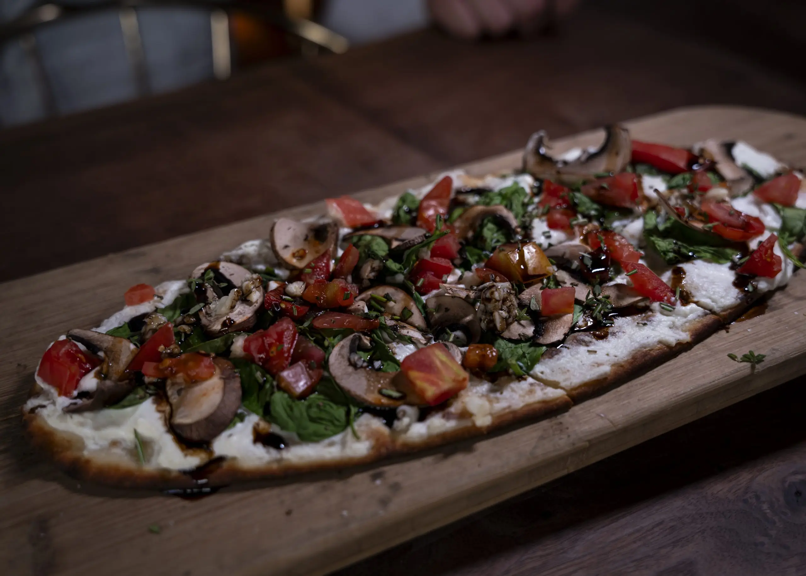 A long rectangular flatbread topped with mushrooms, roasted tomatoes and greens on a wooden board on a dark table.