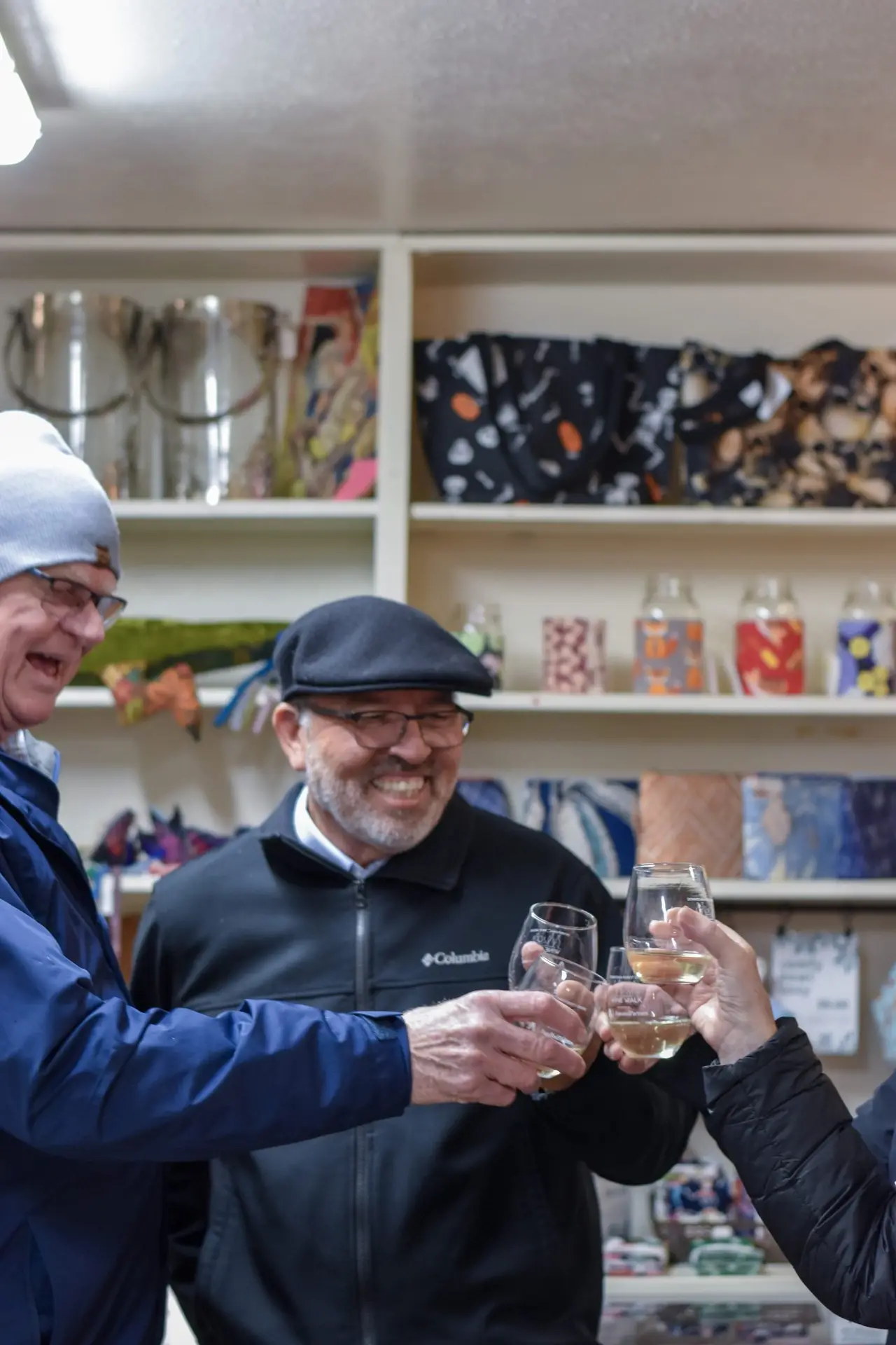 Two men touching wine glasses together inside a shop lined with retail shelving, one in a flat cap.
