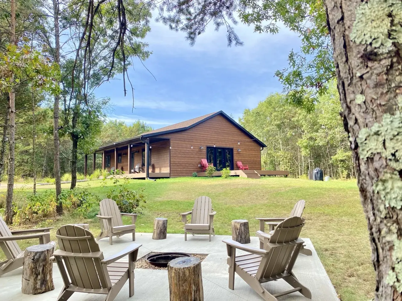 A backyard with a fire pit surrounded by Adirondack chairs on a concrete patio, with a wooden house in the background and a wooded area surrounding the yard.