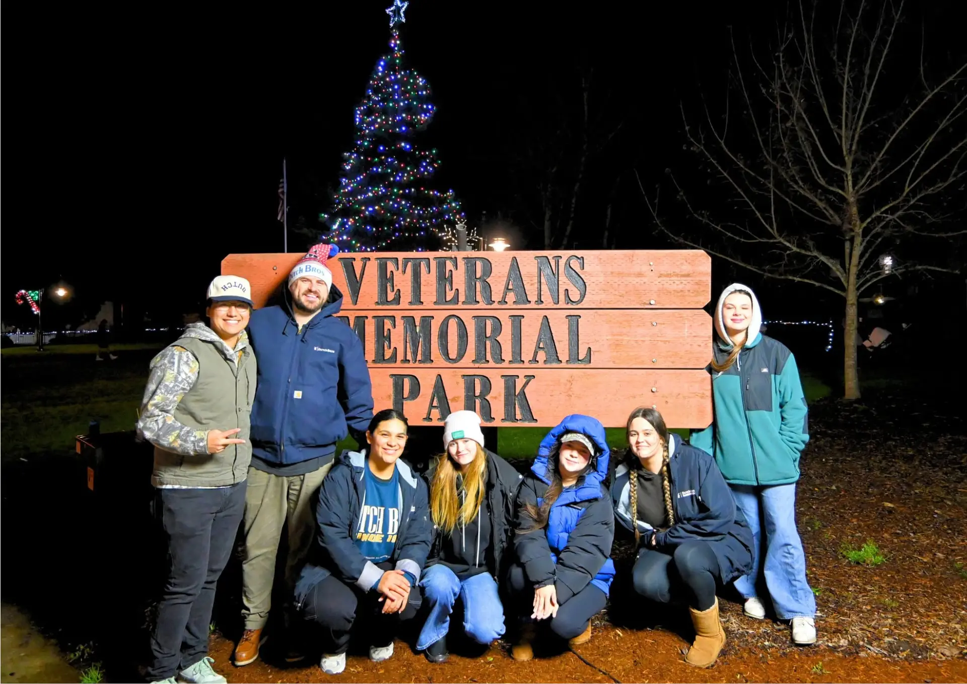 A group of volunteers poses together in front of the Veterans Memorial Park sign and a brightly lit Christmas tree at night.
