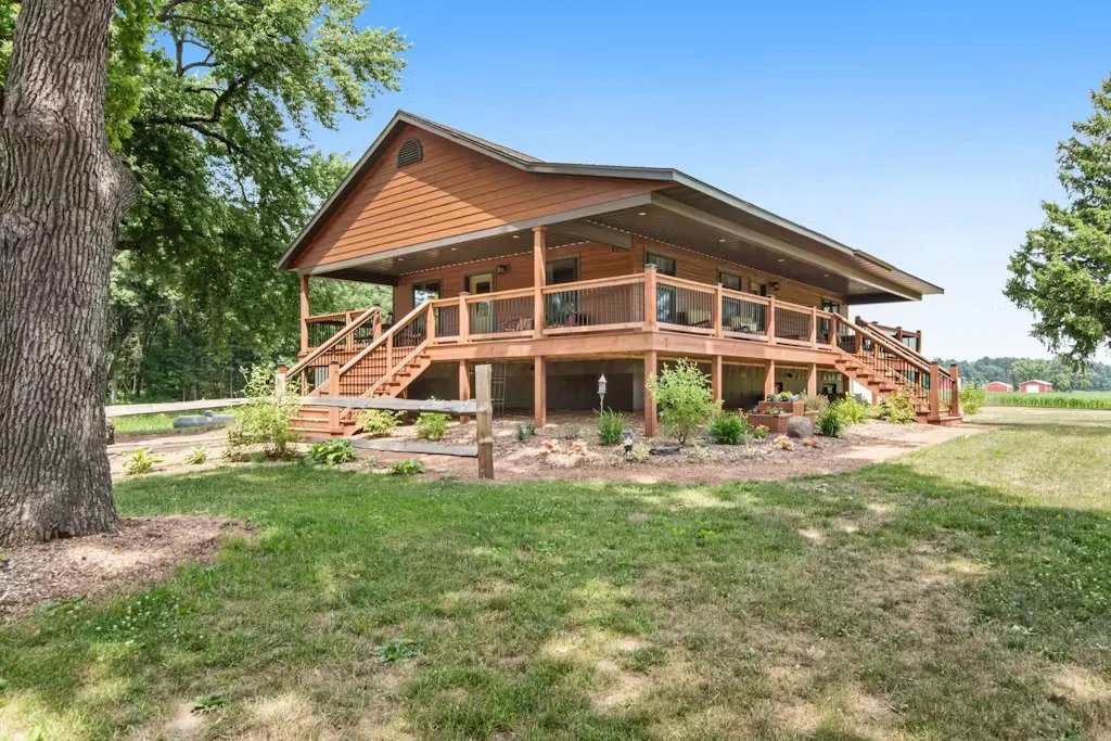Wooden house with a porch and stairs, surrounded by grass and trees under a blue sky.