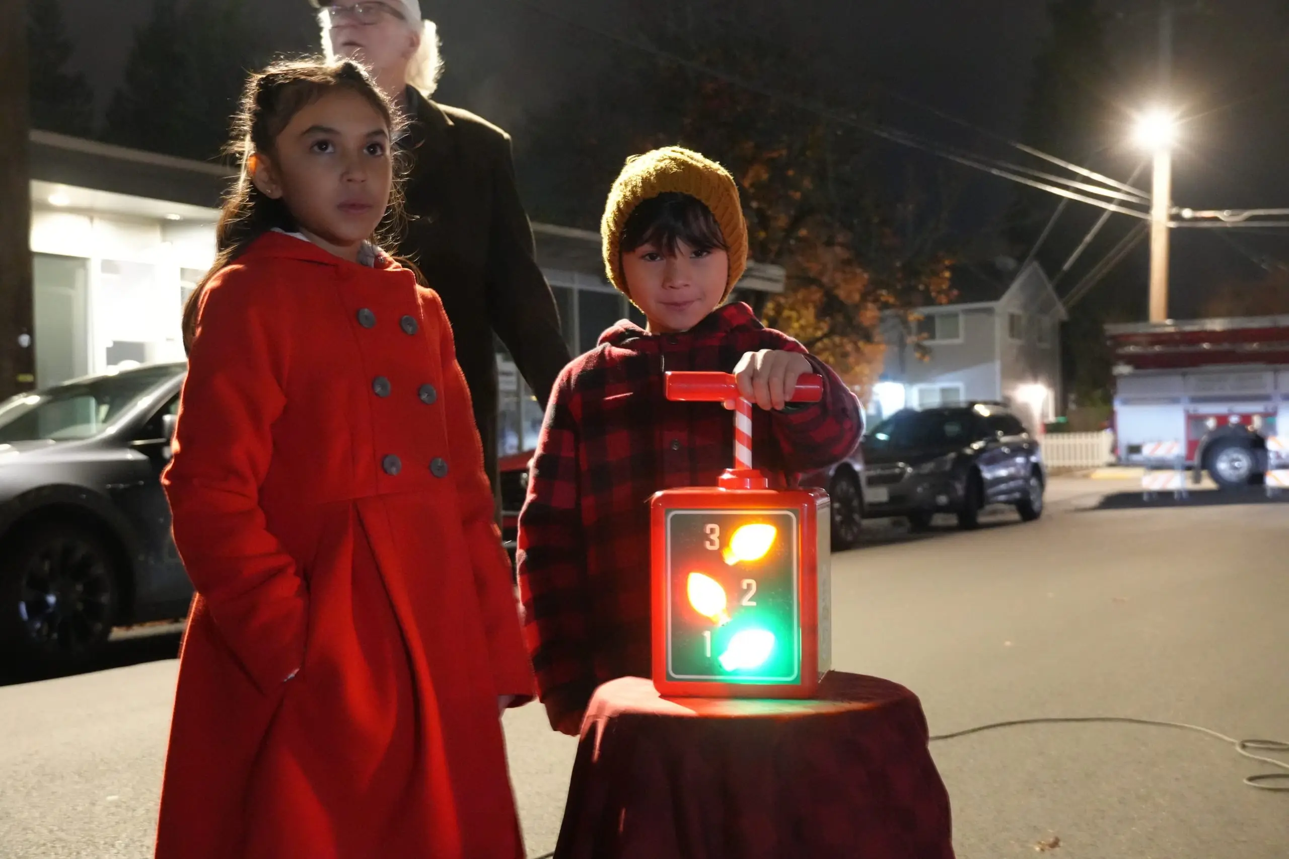 Two children in red winter coats standing on a street at a night-time community event.