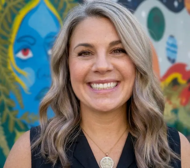 A smiling woman with long fair hair and a round pendant necklace, photographed against a brightly painted mural.
