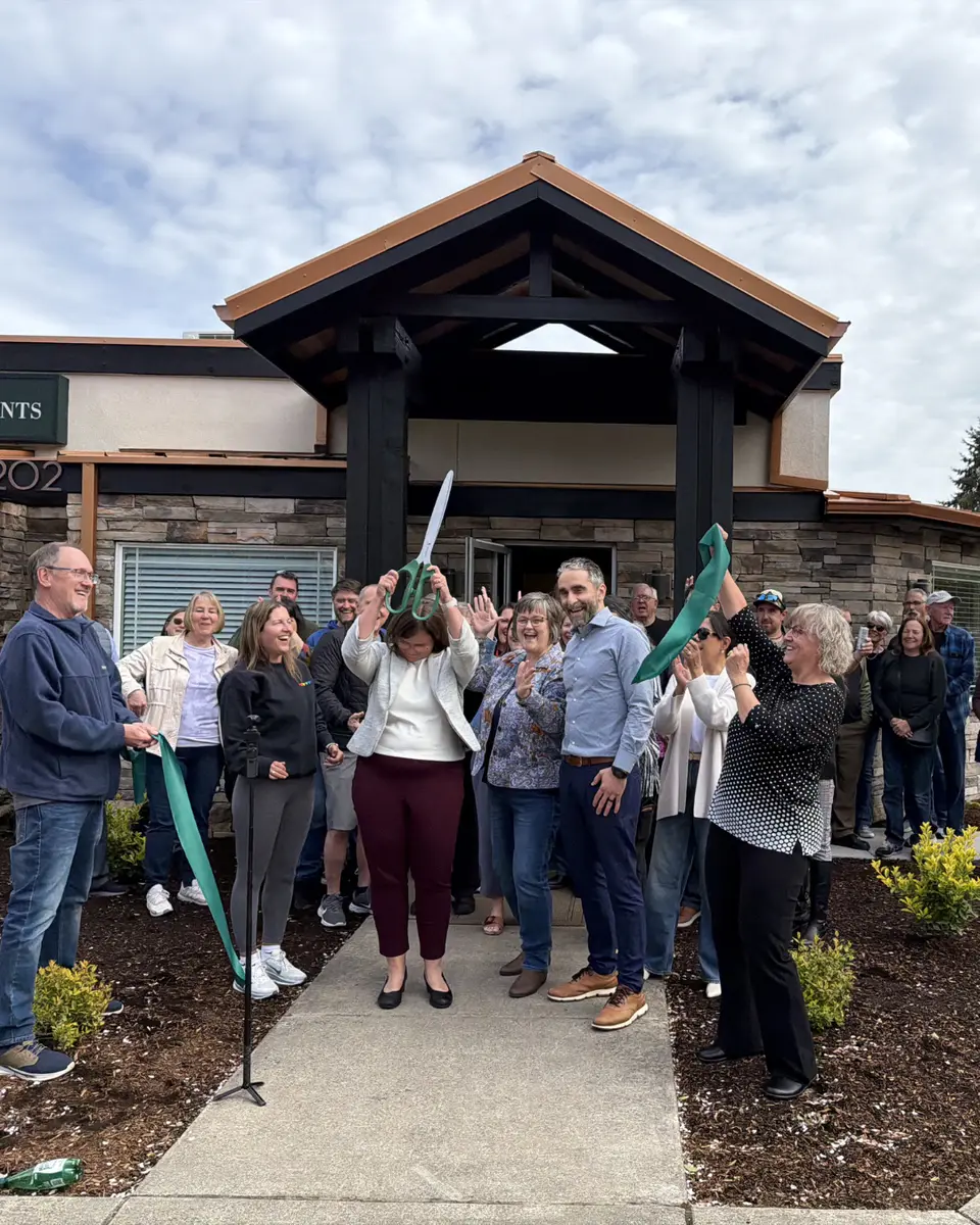 A woman raises oversized scissors after cutting a green ribbon as a crowd of community members cheers and applauds outside a local business.