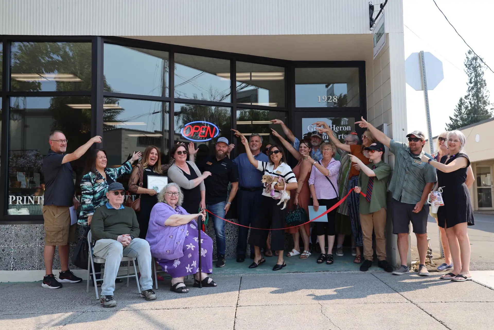 About sixteen people standing outside a single-storey commercial building holding a ribbon, one pair with oversized ceremonial scissors.