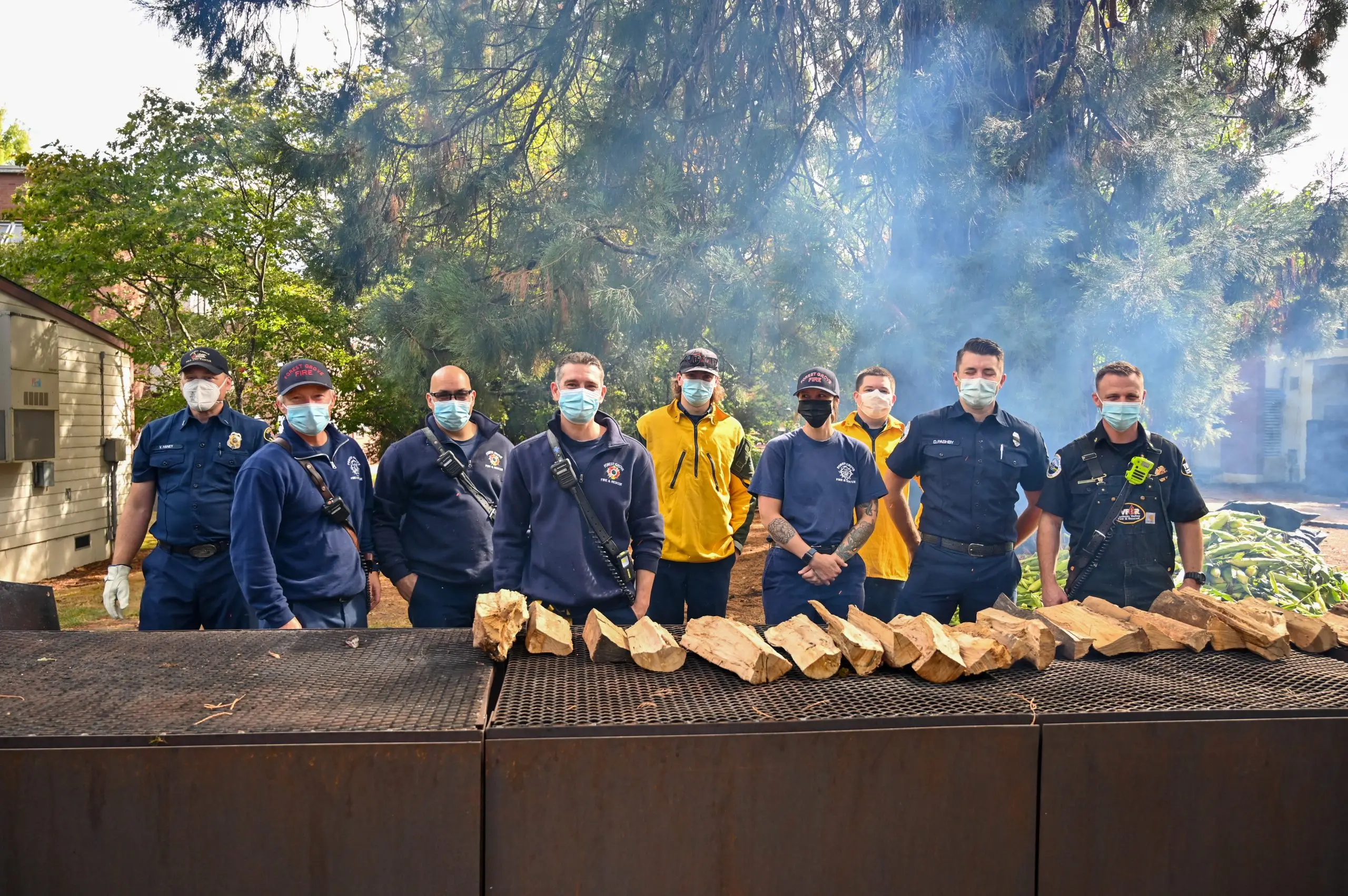 Nine uniformed firefighters and first responders standing in a line behind a long steel grill laid with roasting corn, wood smoke rising.