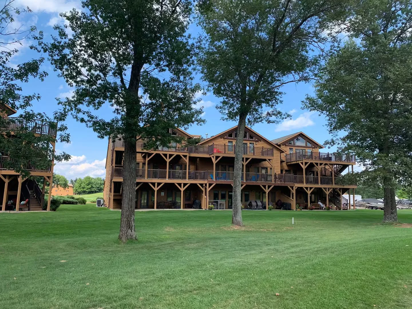A large wooden multi-story building with balconies, surrounded by green grass and trees on a clear day.