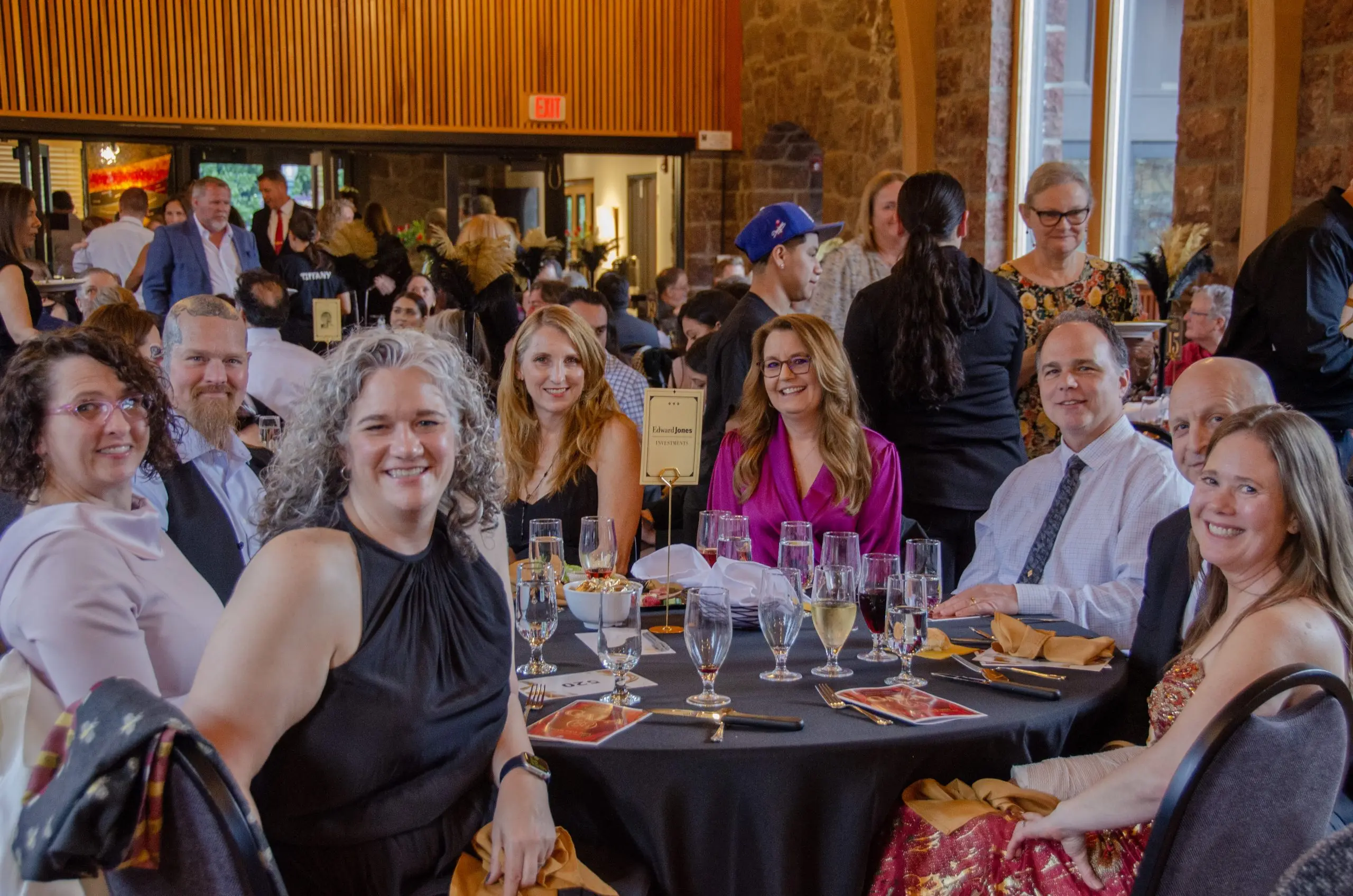 Guests at a round table in evening dress turned toward the camera, a crowd of other attendees behind them.