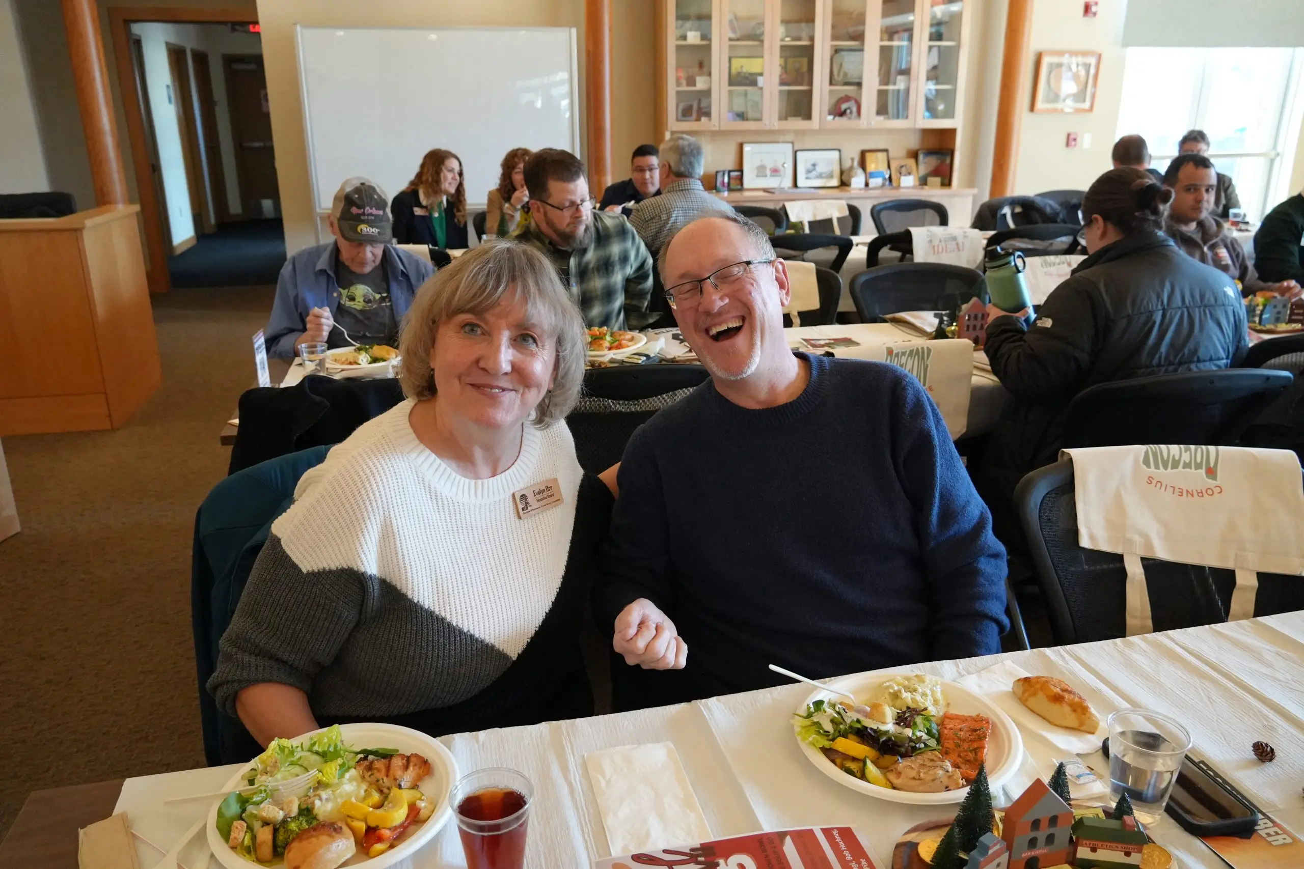 A woman and a man seated side by side at a chamber luncheon table, both laughing, plates of food in front of them.