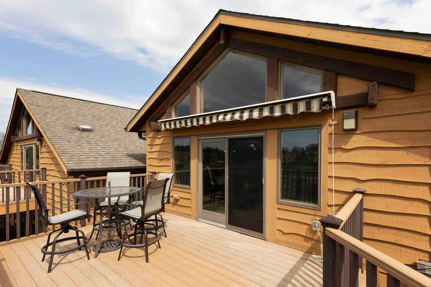 Wooden deck with outdoor furniture, including a round table and four chairs, attached to a house with large windows, sliding door, striped awning, and neighboring house in the background under partly cloudy sky.