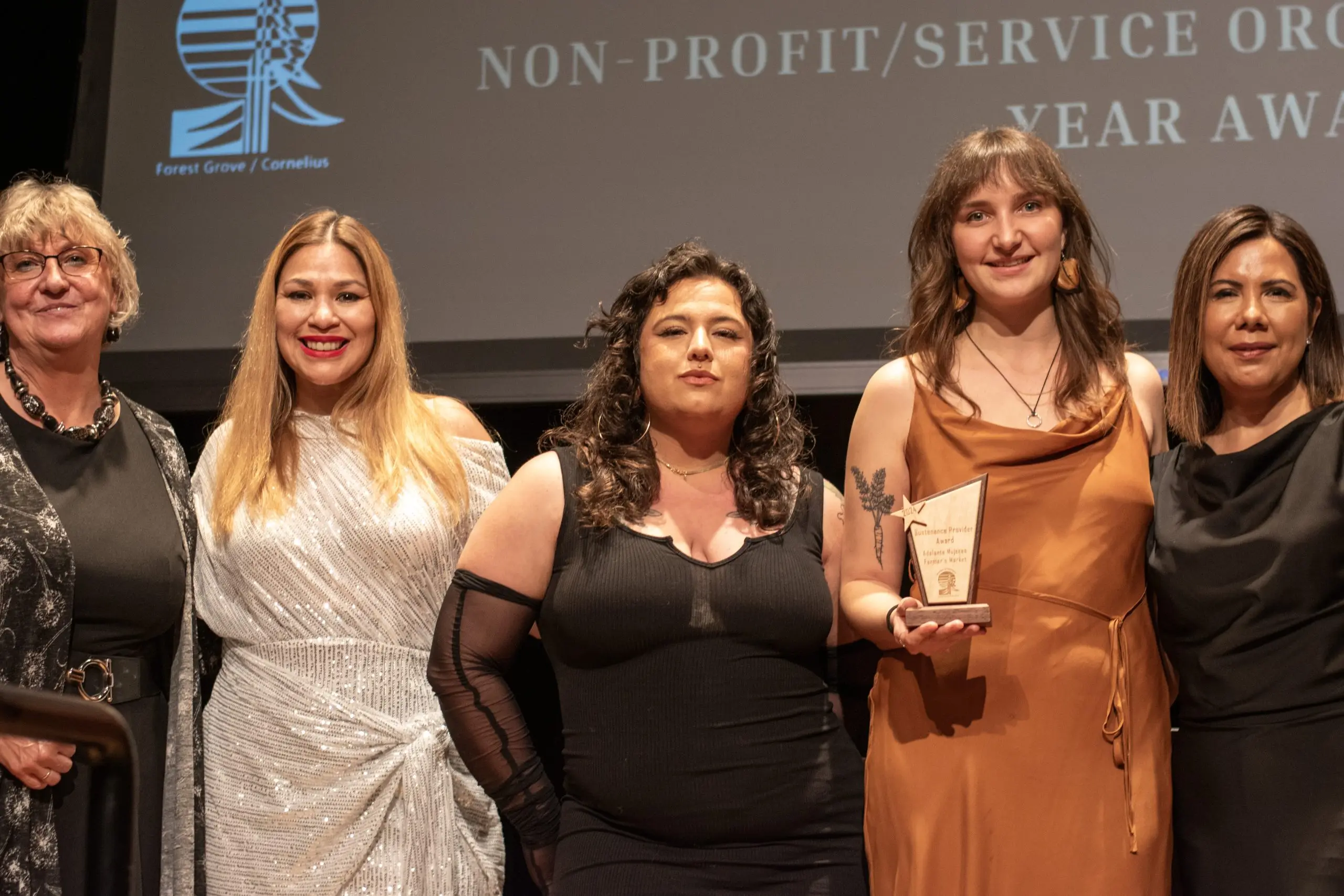 Five women standing shoulder to shoulder on a stage, the second from right holding a small wooden award, in front of a projected slide reading Non-Profit / Service Organization of the Year Award beside the chamber logo.