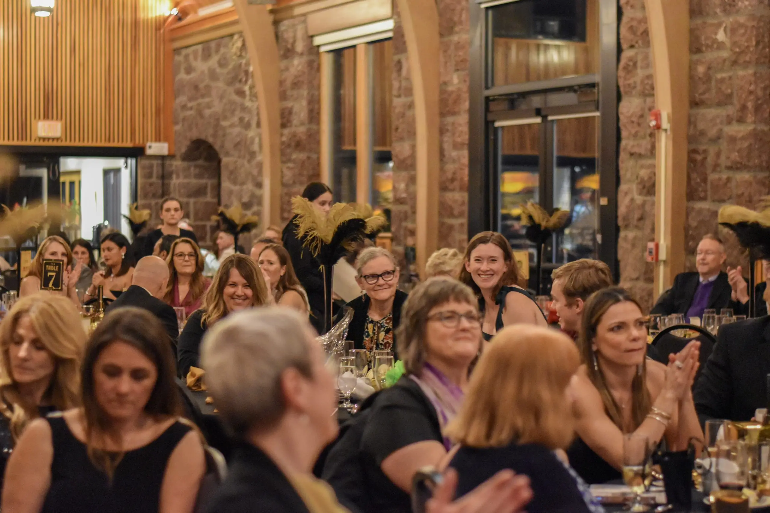 A stone hall packed with guests at round tables, many applauding, tall gold centerpieces throughout.