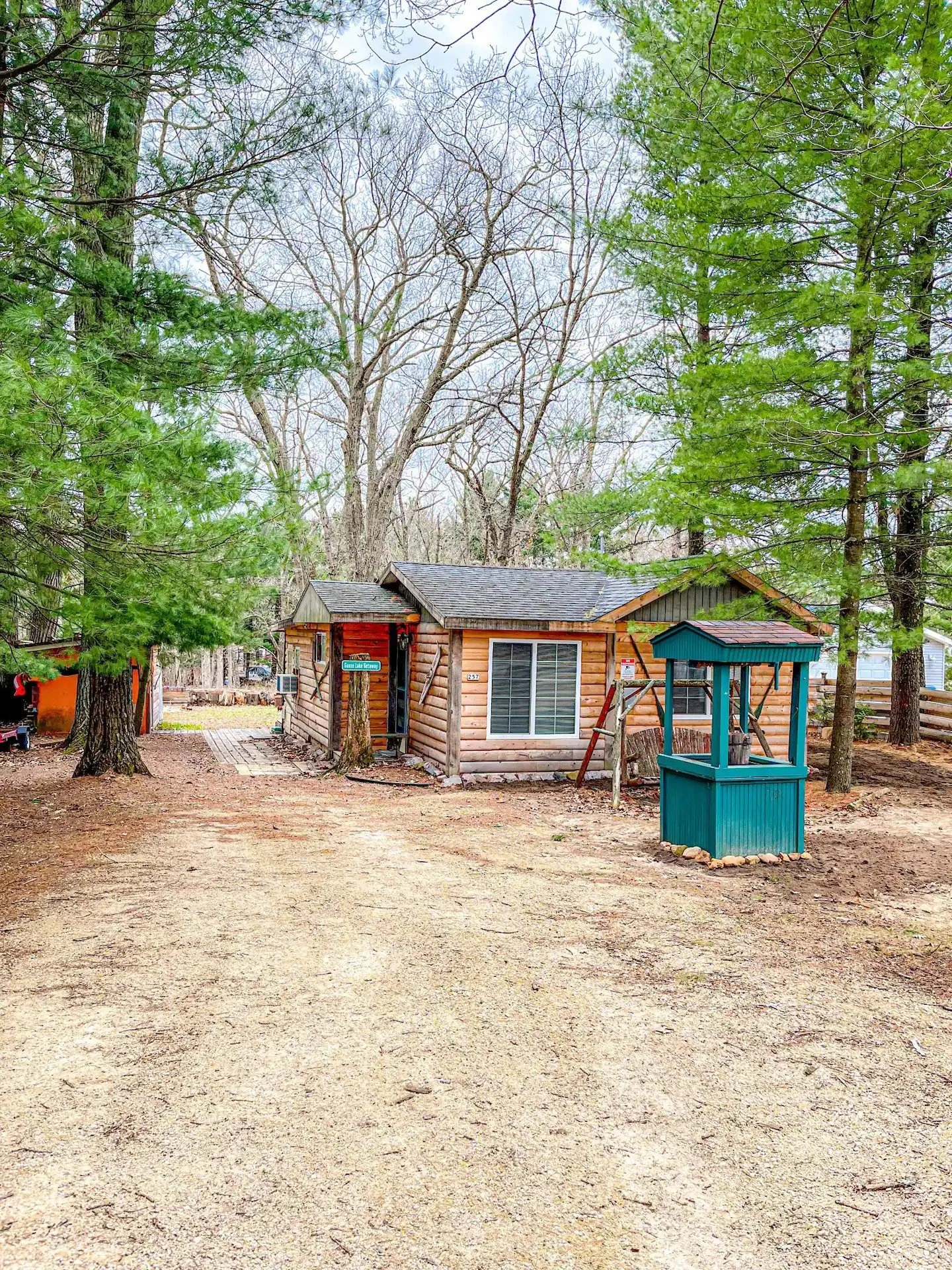A small wooden cabin with a gray shingled roof, surrounded by trees, with a dirt driveway in front. There is a small well or storage shed painted turquoise nearby and a signpost at the front of the cabin.