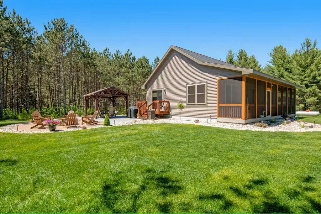 A backyard with a well-maintained green lawn, a house with a screened porch, and a wooden deck with outdoor furniture under a pergola, surrounded by trees and a clear blue sky.