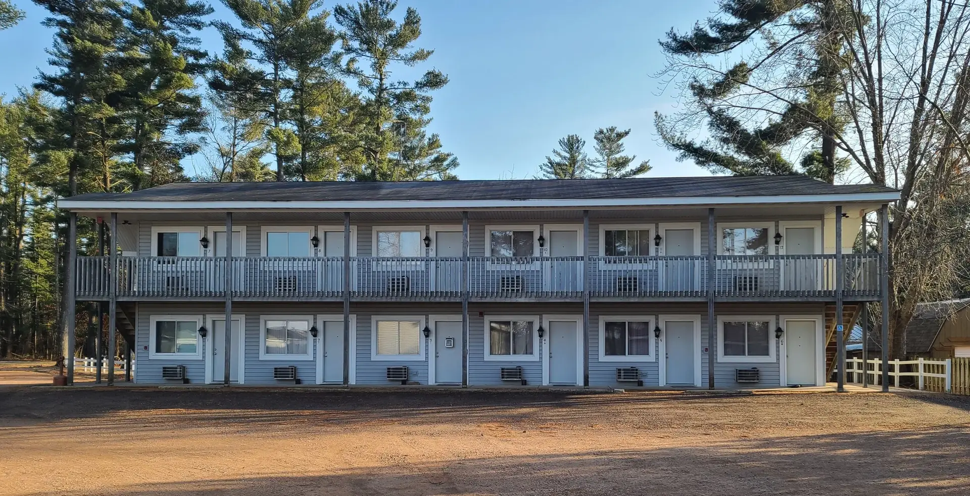 A two-story motel with multiple doors and windows, surrounded by trees, with a dirt parking area in front.