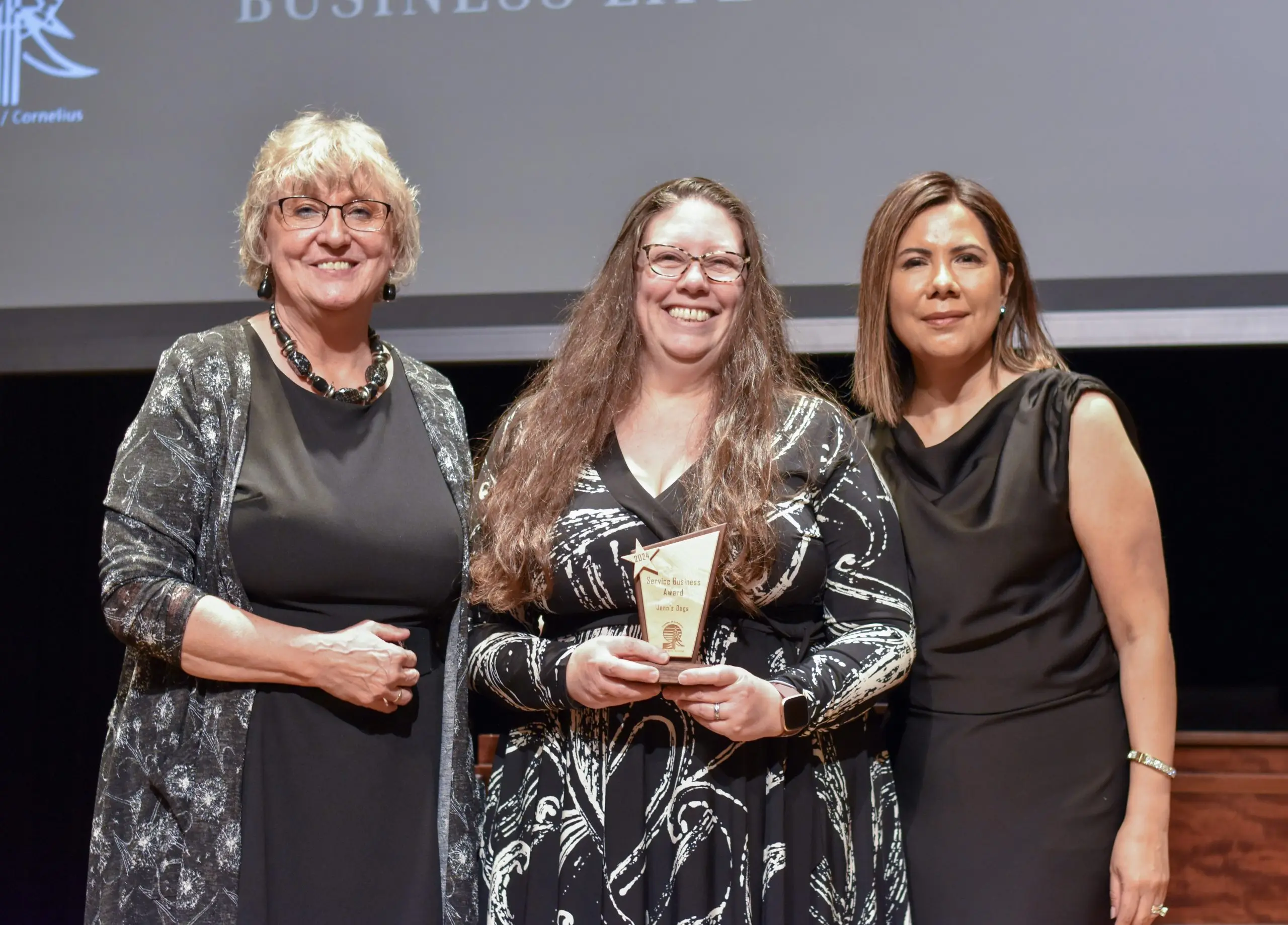 Three women on stage, the one in the center holding a clear glass trophy and beaming, a projection screen behind them.