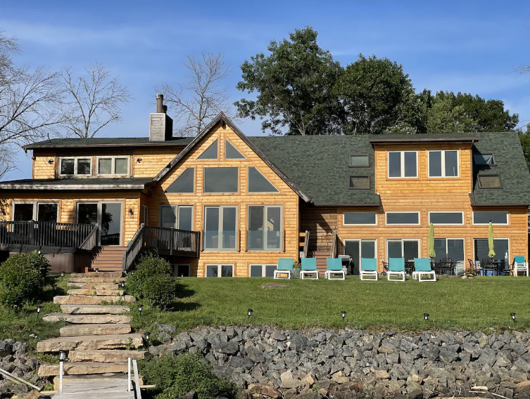 Large house with wooden siding, multiple large windows, and a green roof, sitting on a grassy yard with outdoor chairs and trees in the background.