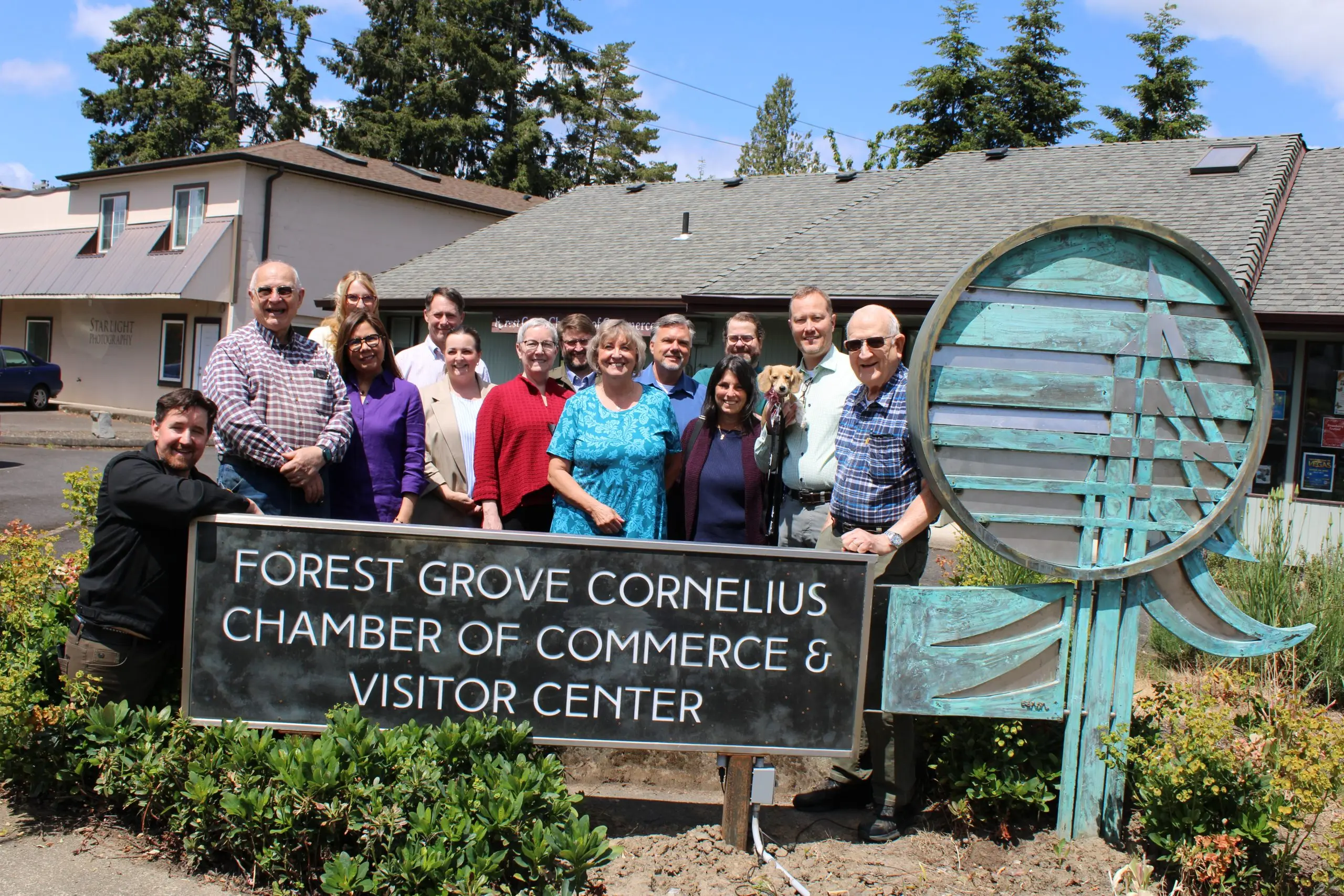 Chamber board members and staff standing together outside the chamber office, a tall verdigris metal sculpture beside them and firs behind.