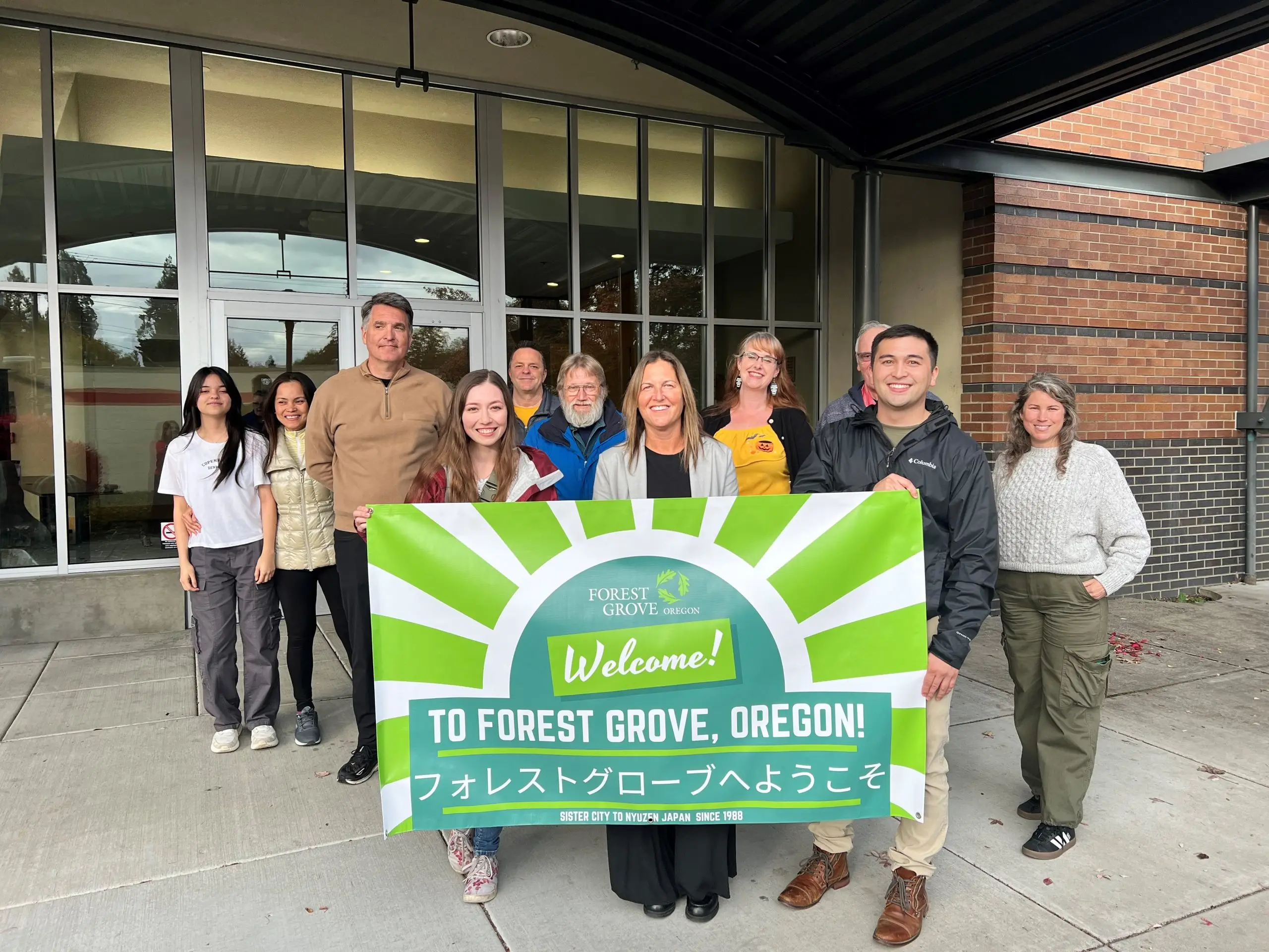 Eleven people standing under the canopy of a glass-fronted civic building holding a green banner with the Forest Grove Oregon mark on it above the word Welcome.