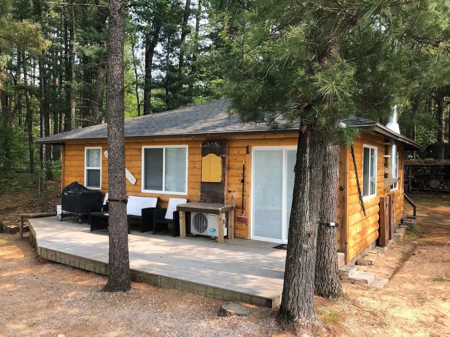 A small wooden cabin in a forest with a wooden deck, outdoor seating, a barbecue grill, and an air conditioning unit.