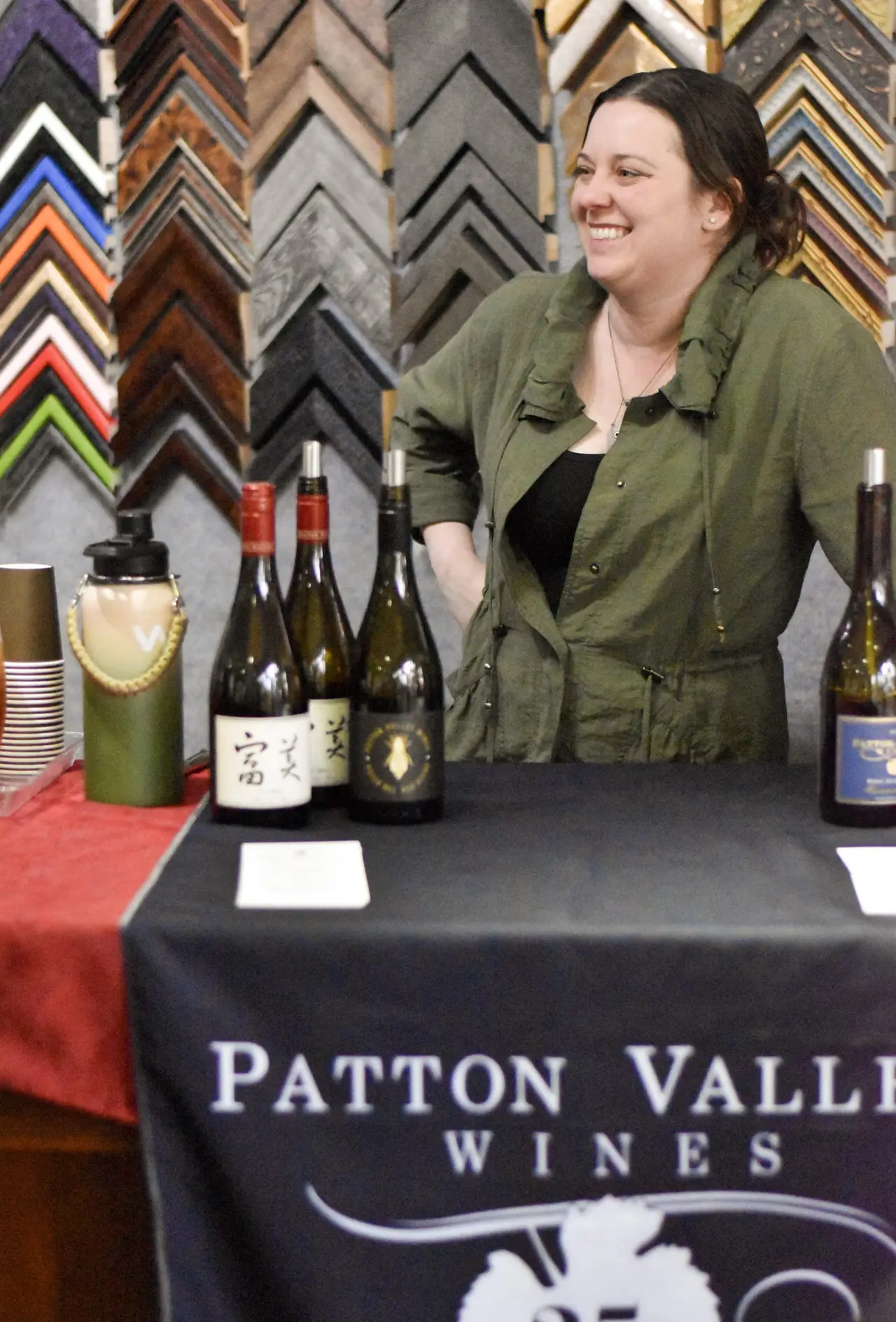 A woman standing behind a pouring table of wine bottles, the wall behind her hung with rows of picture-frame corner samples.
