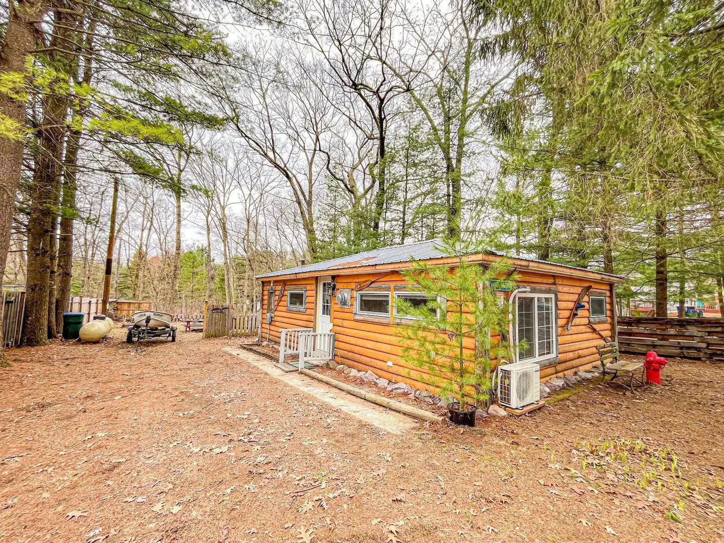 A small log cabin in a wooded backyard with leafless and evergreen trees. The house has a metal roof, multiple windows, and an air conditioning unit. There is a bench, a fire hydrant, a boat, and a propane tank nearby, with a dirt ground and a fence surrounding the area.