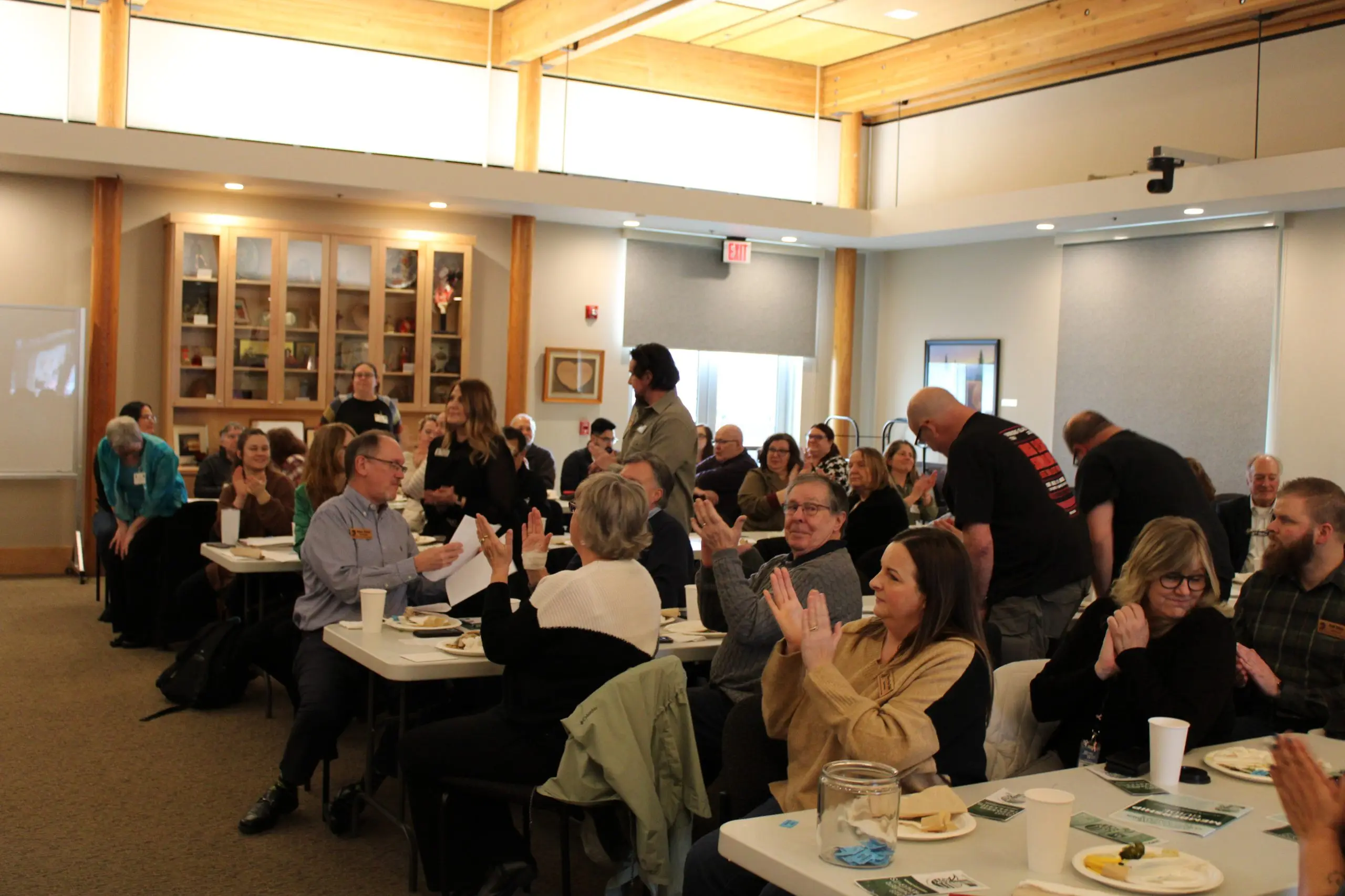 A community room with a warm timber ceiling full of people standing and seated at tables with coffee cups and plates, talking in groups.