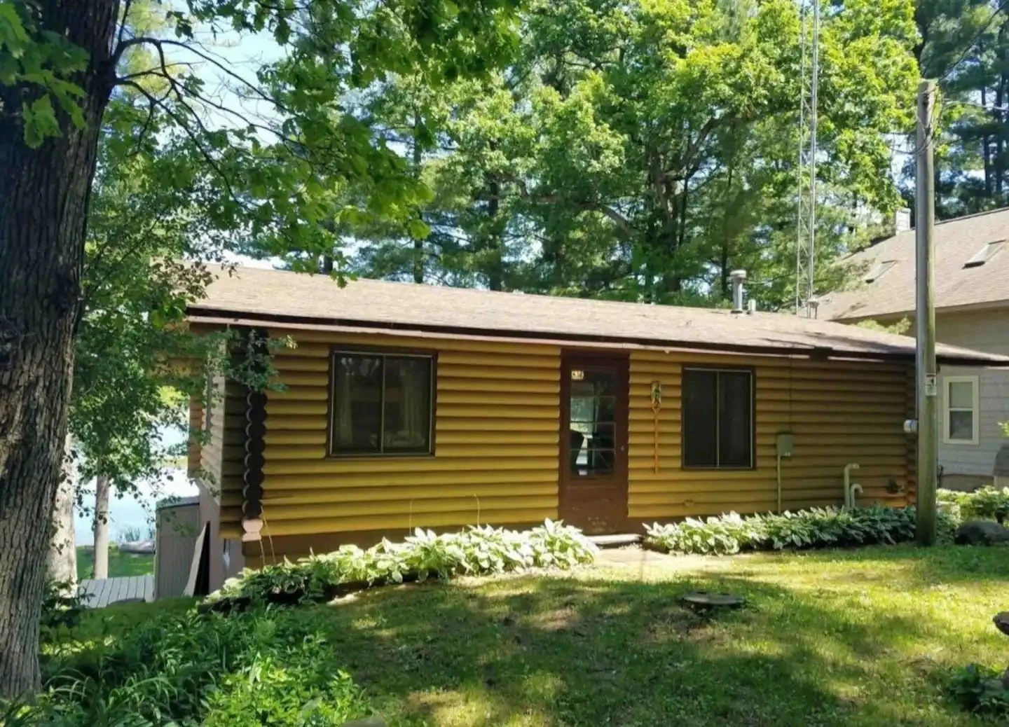 A small brown house with horizontal siding, two windows, and a door, surrounded by green trees and plants, with a yard in the foreground.