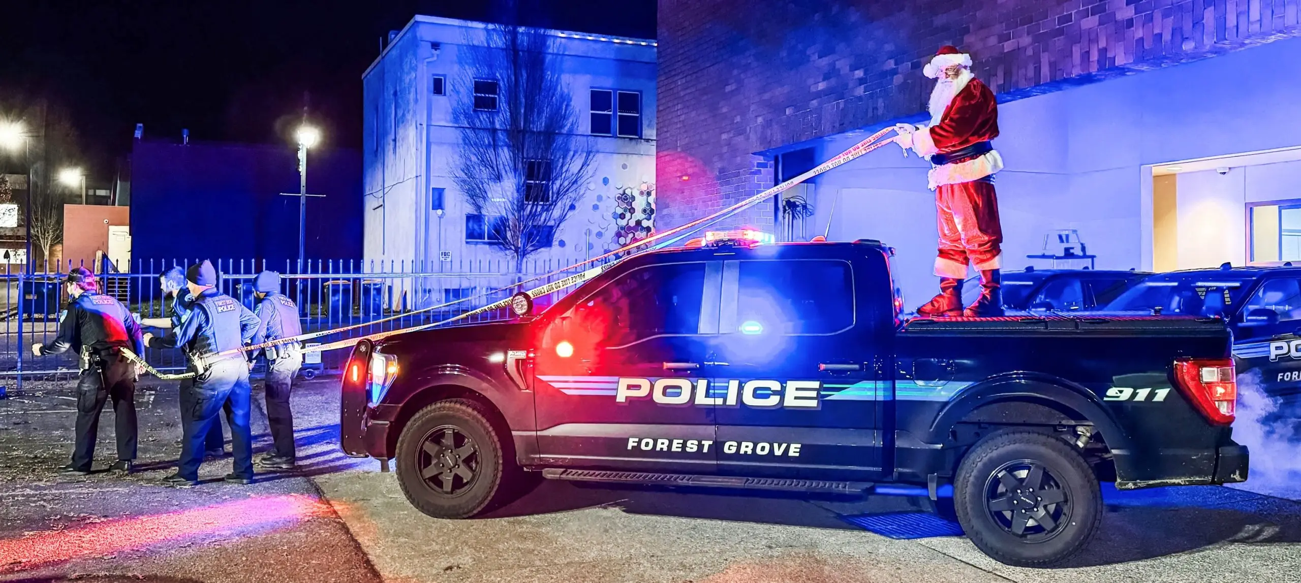Santa Claus standing in the bed of a marked Forest Grove Police pickup truck at night, lit by red and blue emergency lights.