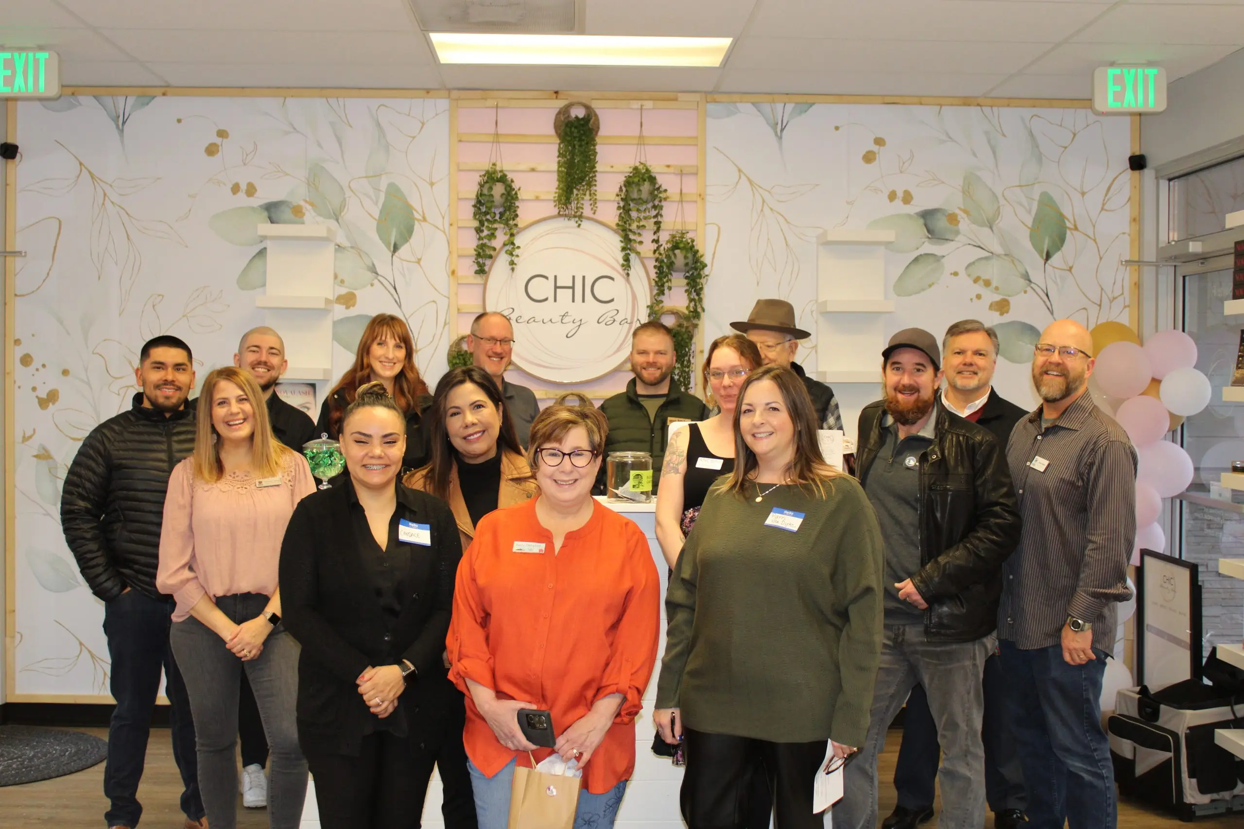 About fifteen people wearing name badges posed together in a bright salon in front of a botanical mural and a hanging sign reading CHIC, pink and white balloons at the right.