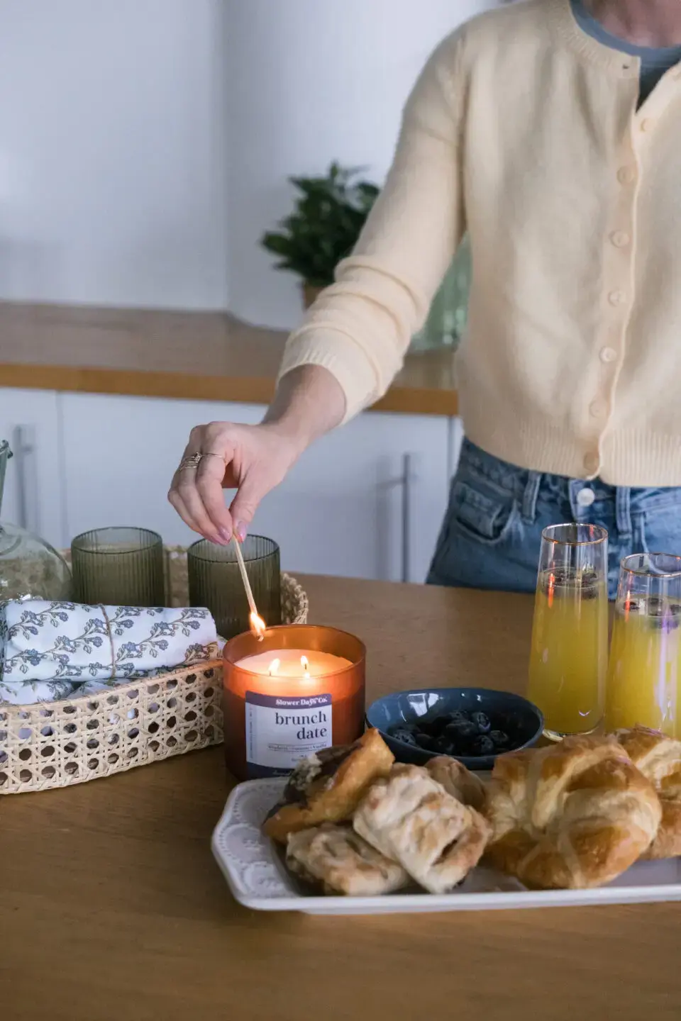 A hand lighting a Slower Days Co. “Brunch Date” candle on a table with pastries, blueberries, and drinks nearby
