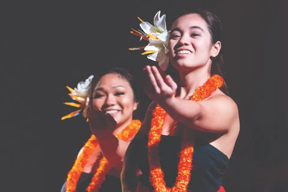 Two young women dancing in Polynesian costume with orange feather leis and white flower headpieces on a dark stage, hands raised mid-gesture.