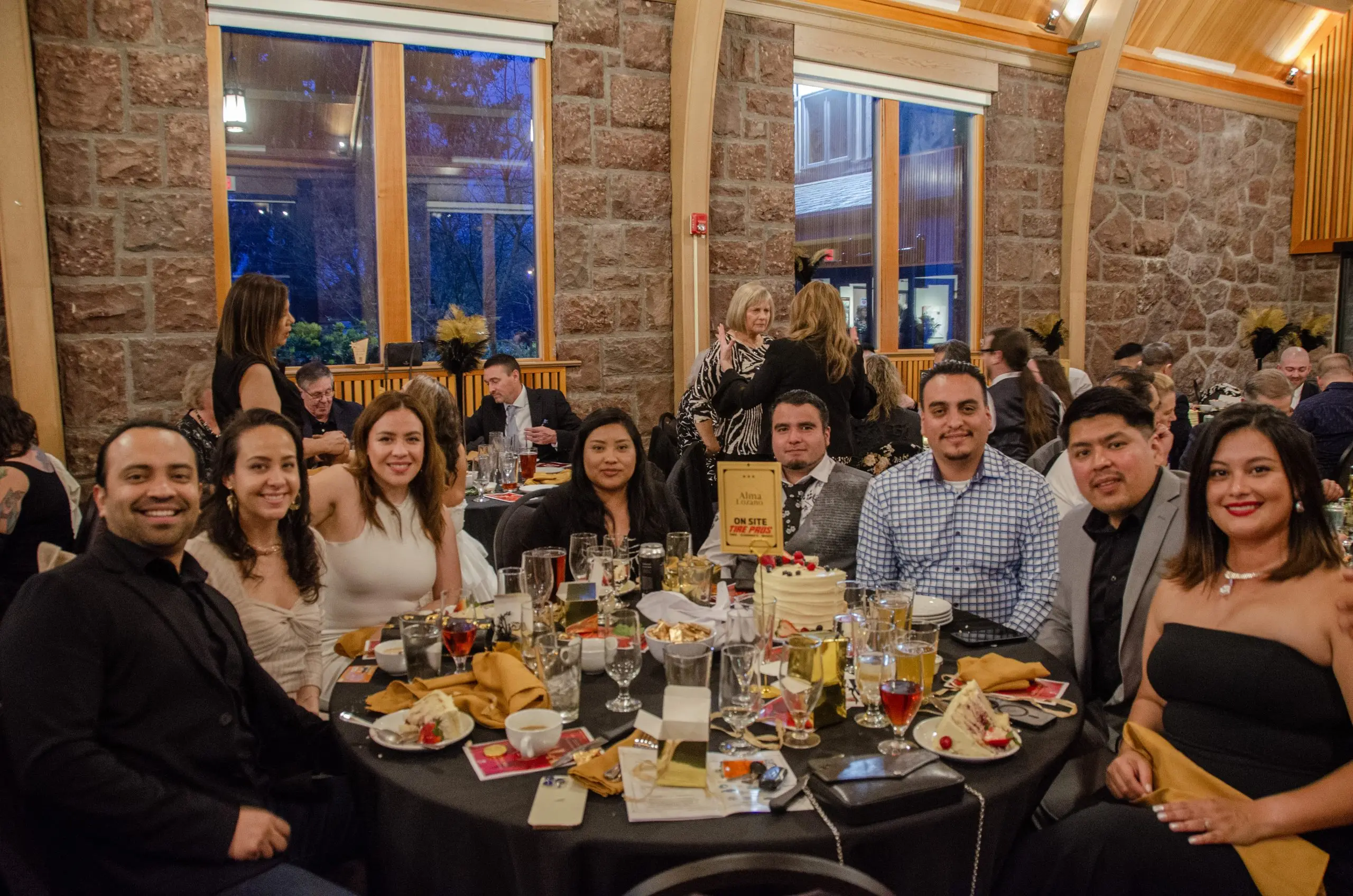 Seven guests in evening dress around a black-linen round table with gold napkins and plated food, all turned to the camera.