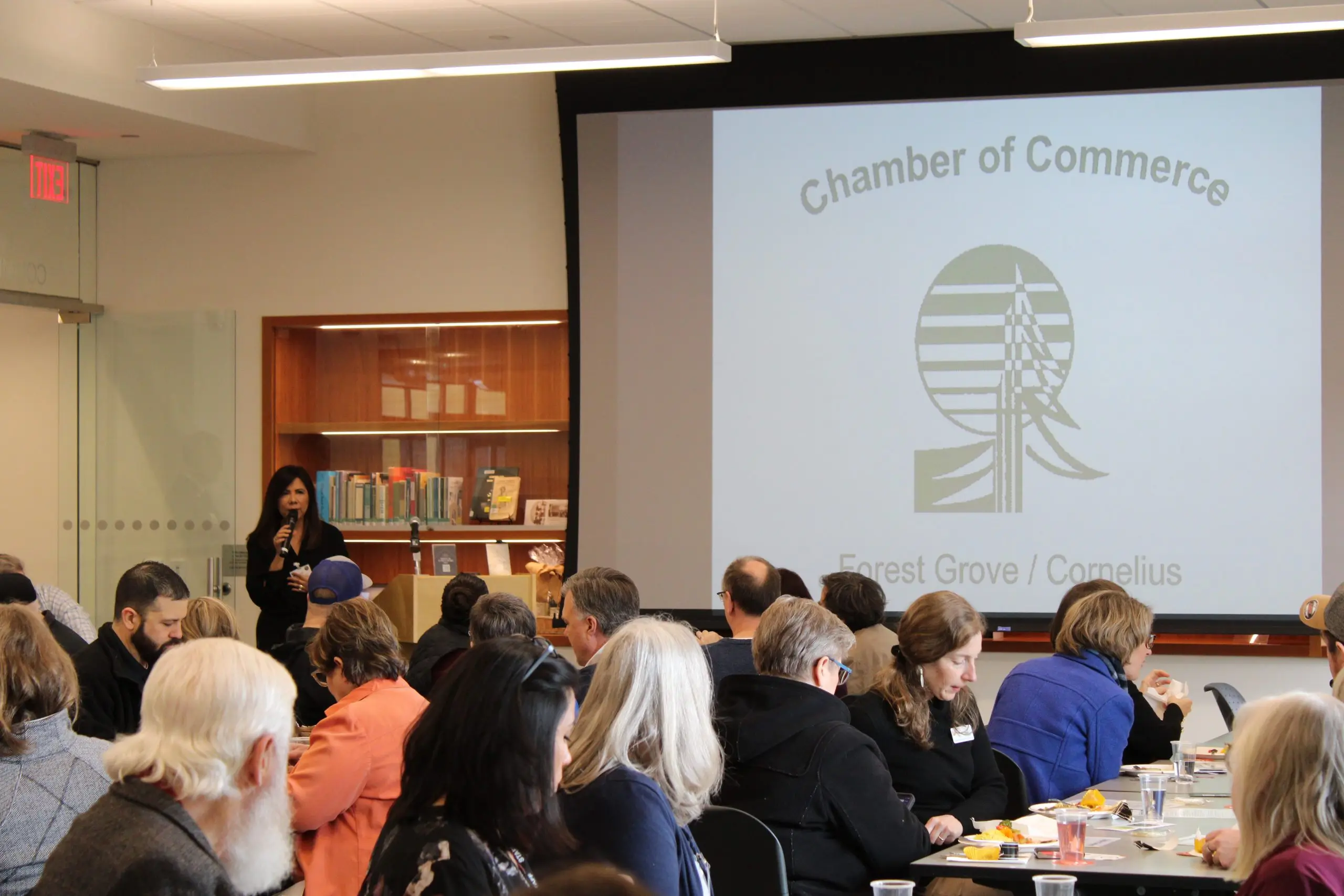 An audience seated at tables in a library meeting room facing a woman with a microphone, the projection screen beside her carrying the chamber tree logo and the words Forest Grove / Cornelius.