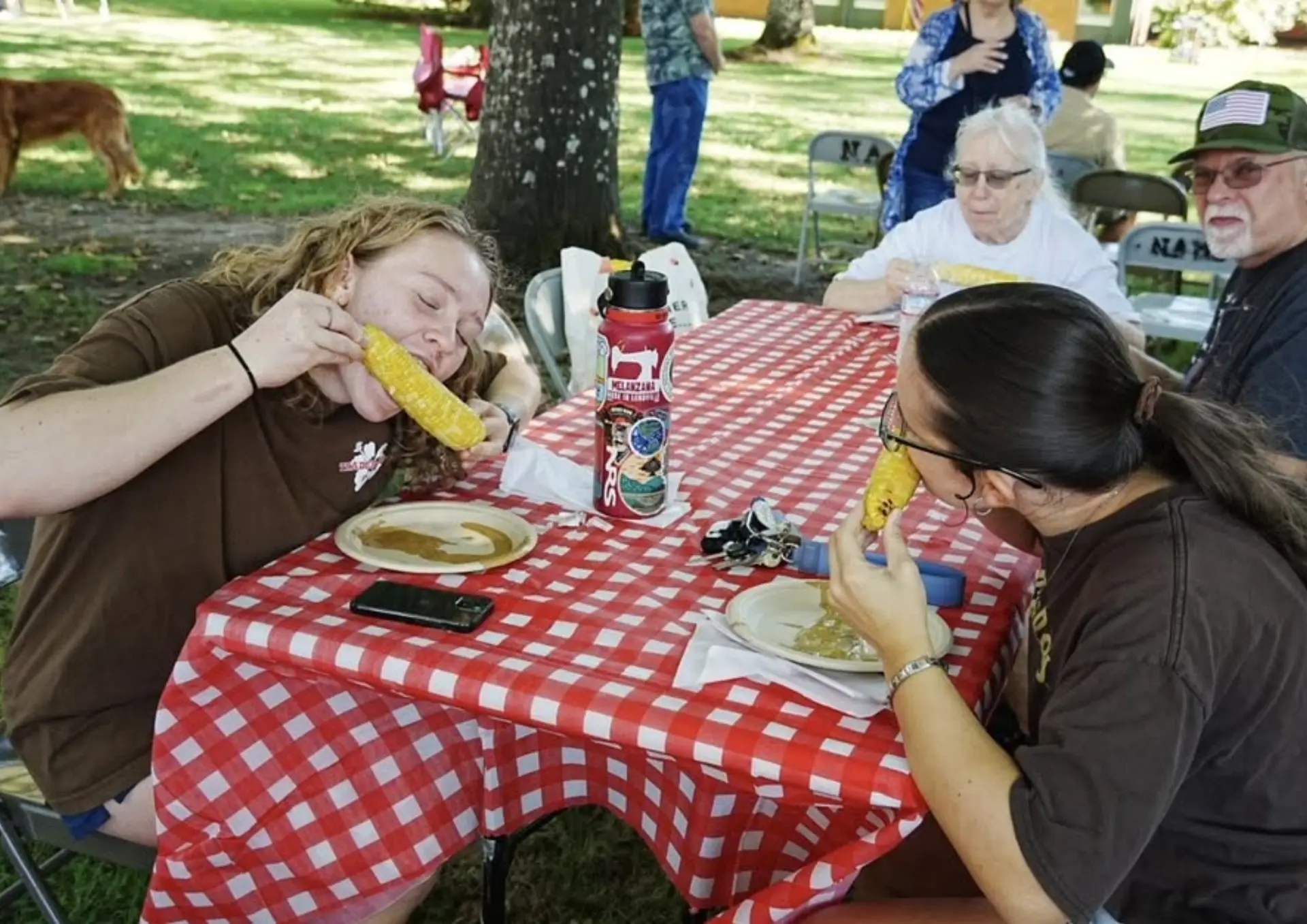 Community members enjoy fresh corn on the cob together at an outdoor picnic table covered with a red-and-white checkered tablecloth.