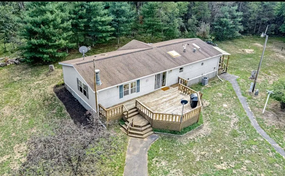 A single-story house with a wooden deck on the backyard, surrounded by grass and trees, with a satellite dish on the roof and a pathway leading to the deck.