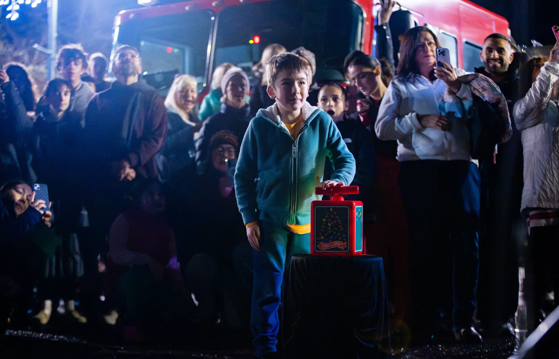 child in coat pushing down on box lever to turn on christmas lights with crowd in background.