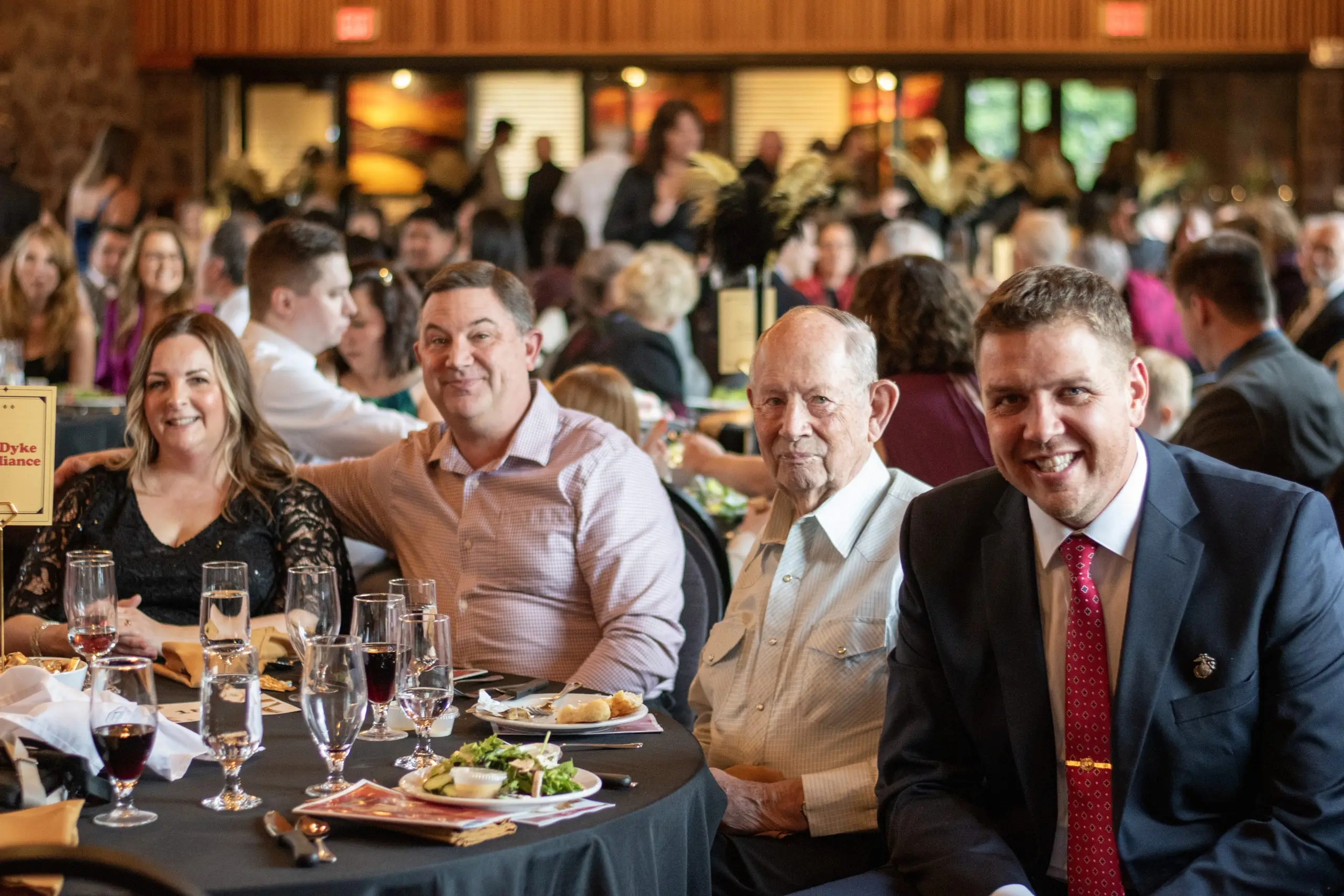 Four guests at a round table in evening dress turned to the camera and laughing, plated dinners in front of them and a full hall behind.