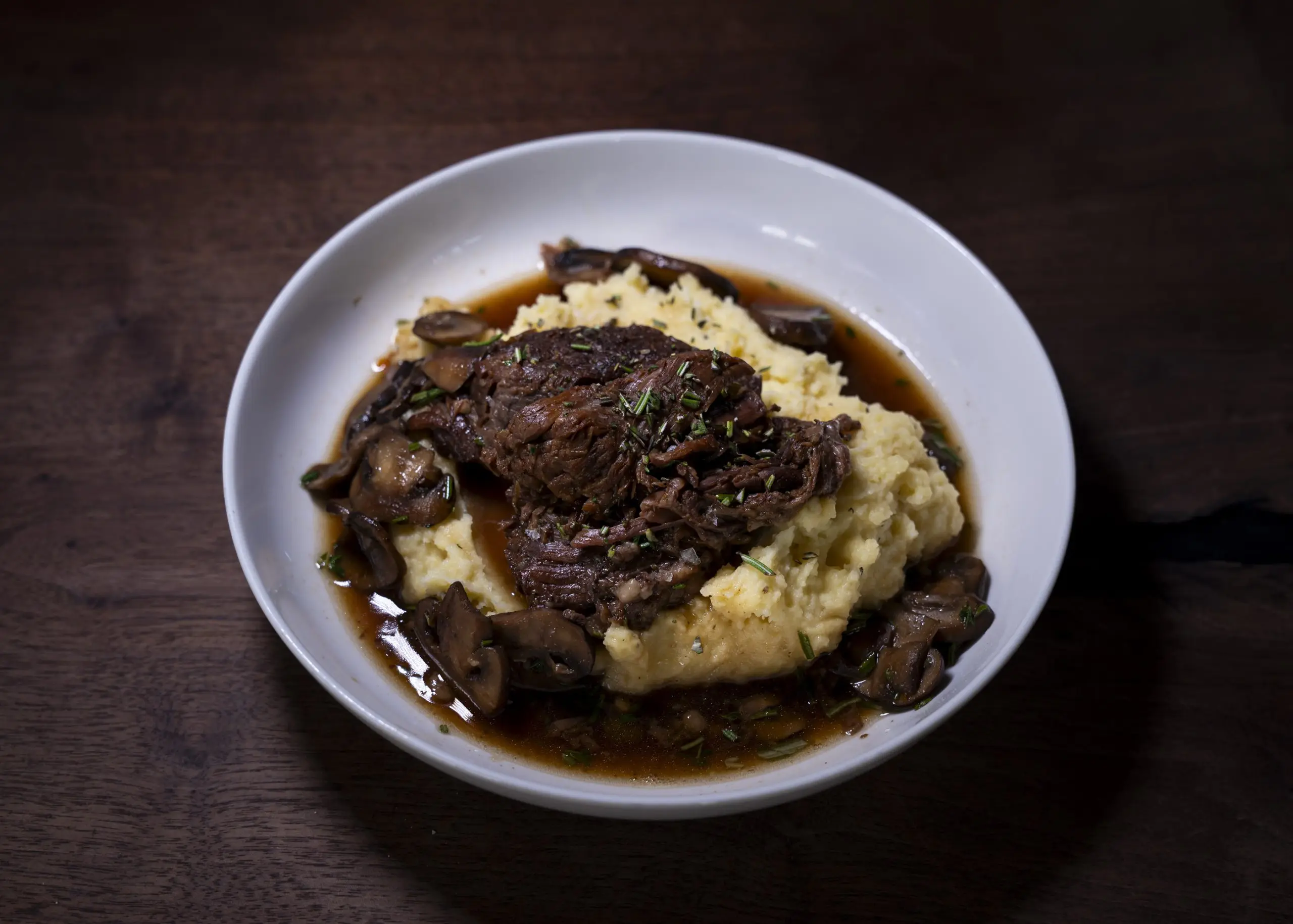 Braised short rib in dark jus over mashed potato in a white bowl on a dark wooden table.