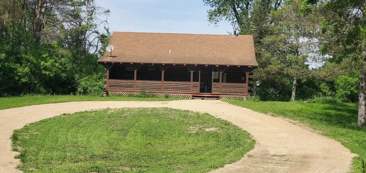 A wooden house with a gabled roof and front porch, surrounded by trees and a gravel driveway.