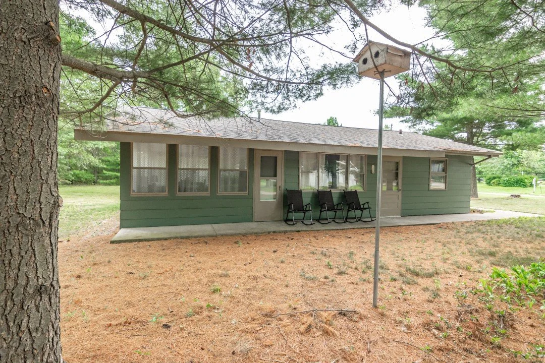 A small green house with multiple windows, a concrete porch, and a gray door, surrounded by pine trees; a birdhouse on a pole and three black lawn chairs outside.