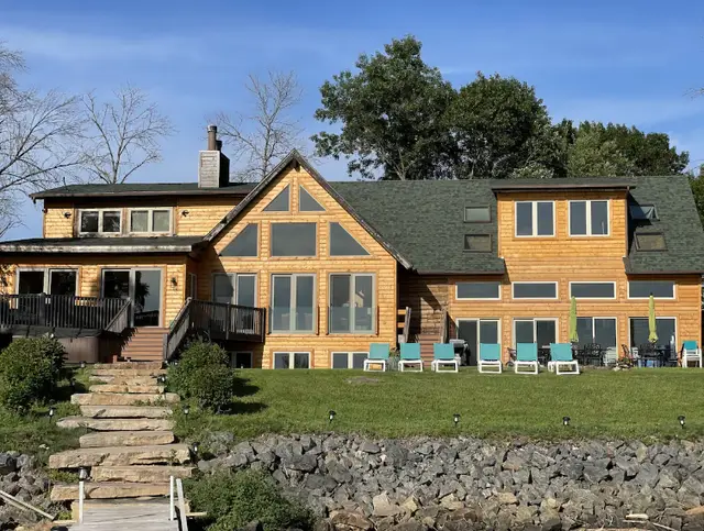 Large house with wooden siding, multiple large windows, and a green roof, sitting on a grassy yard with outdoor chairs and trees in the background.