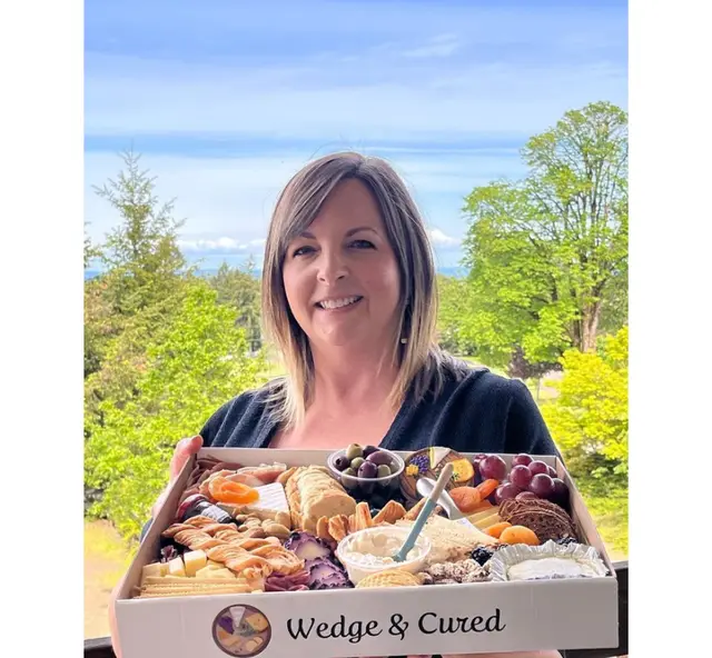 A smiling woman holding out a large white catering box lettered Wedge and Cured, packed with cheese, fruit and cured meats, trees and open sky behind her.