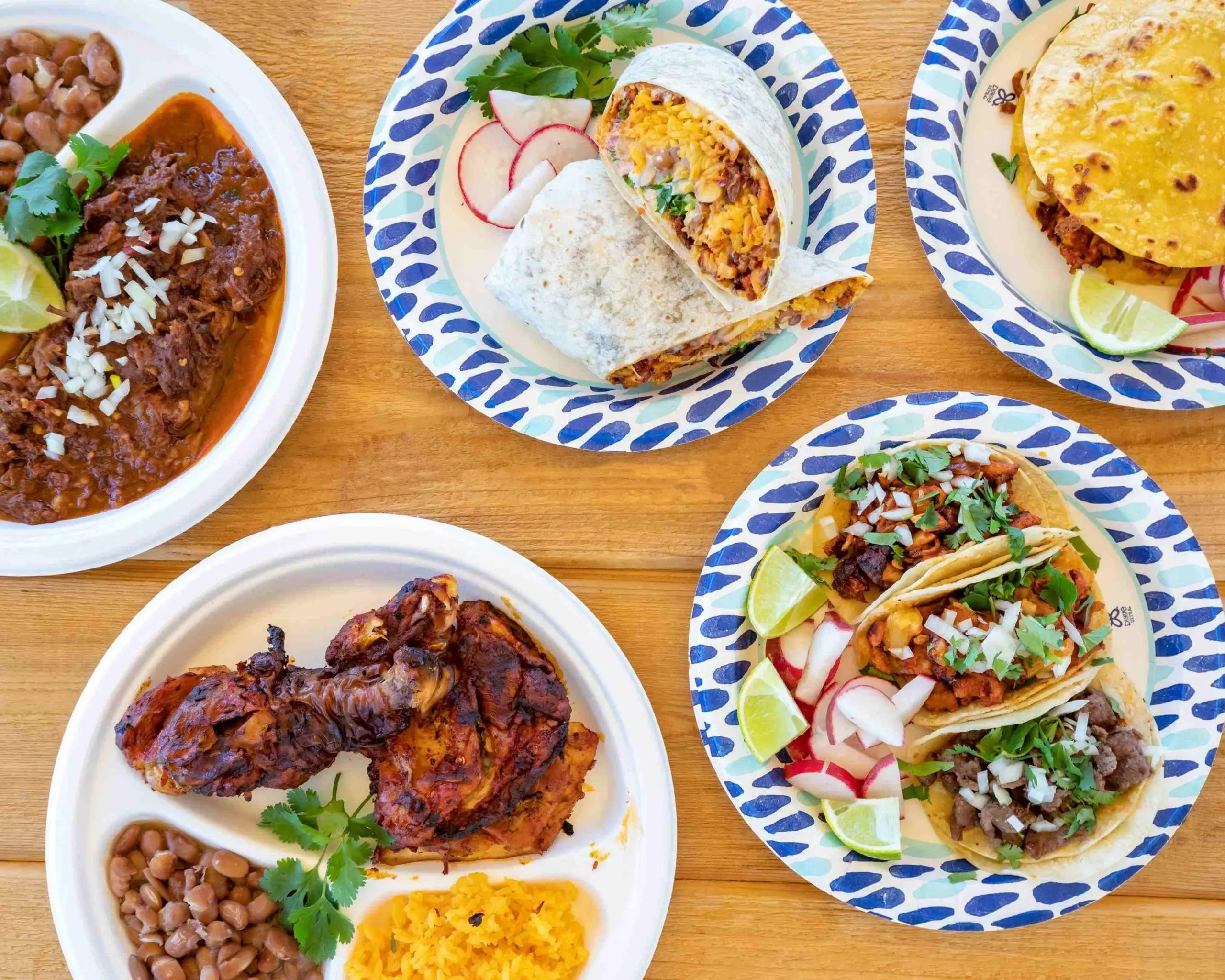 An overhead shot of a Mexican meal on a wooden table: a bowl of beans, plates of rice with radish and cilantro, three tacos on a patterned plate, a quesadilla and grilled meat with corn.