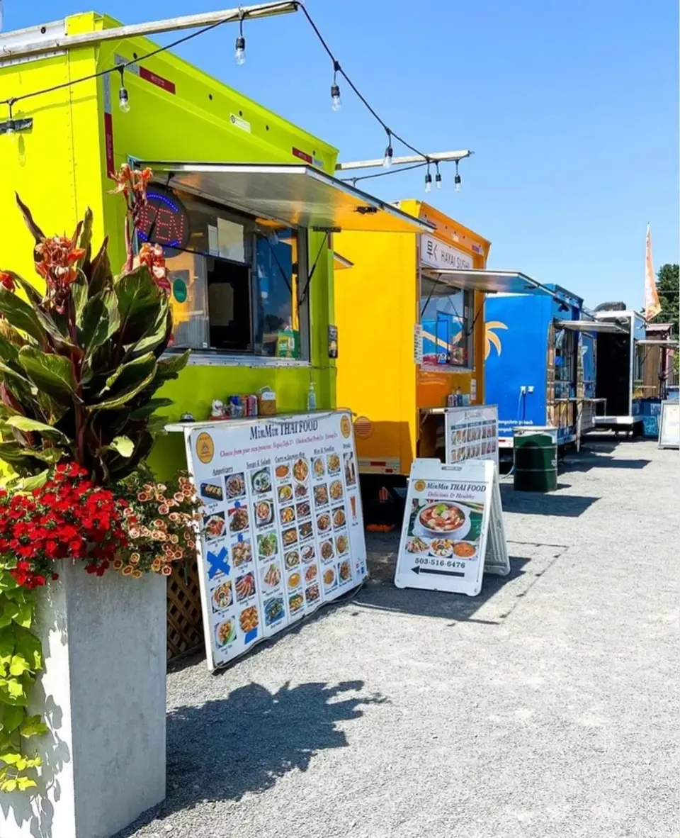 A row of brightly painted food carts under a string of lights, with menu boards and a planter of red flowers in front of them.