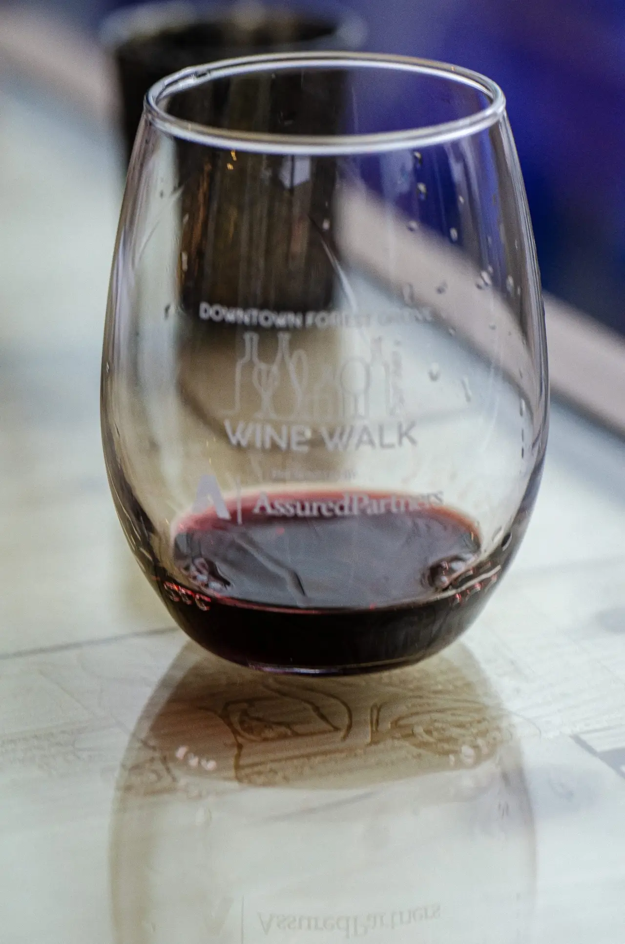 A stemless wine glass of red wine on a white tablecloth, etched Downtown Forest Grove Wine Walk with a sponsor logo beneath the lettering.