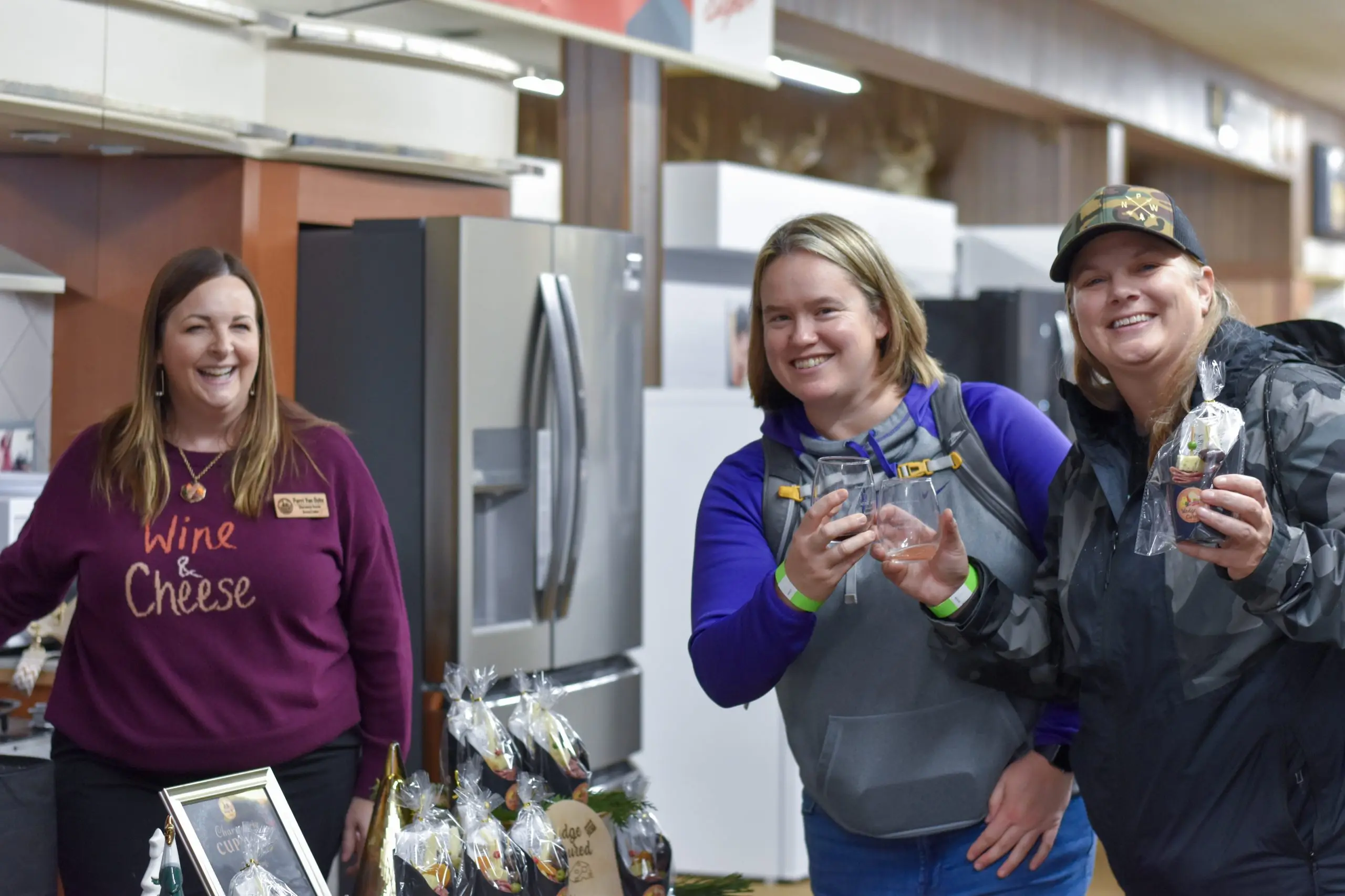 Three women holding tasting glasses and wrapped tasting packages, laughing, the one at left in a Wine and Cheese sweatshirt.
