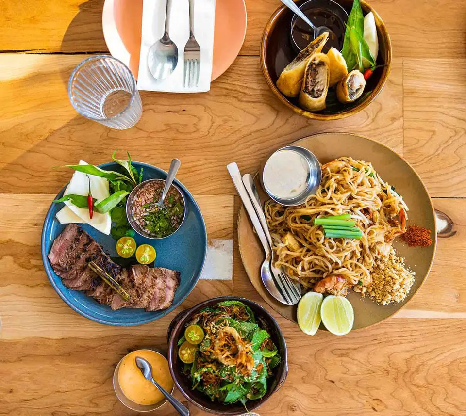 An overhead flat lay of a restaurant table on pale wood: sliced steak with chimichurri, stir-fried noodles with prawns, fried spring rolls, dressed greens and a glass tumbler.